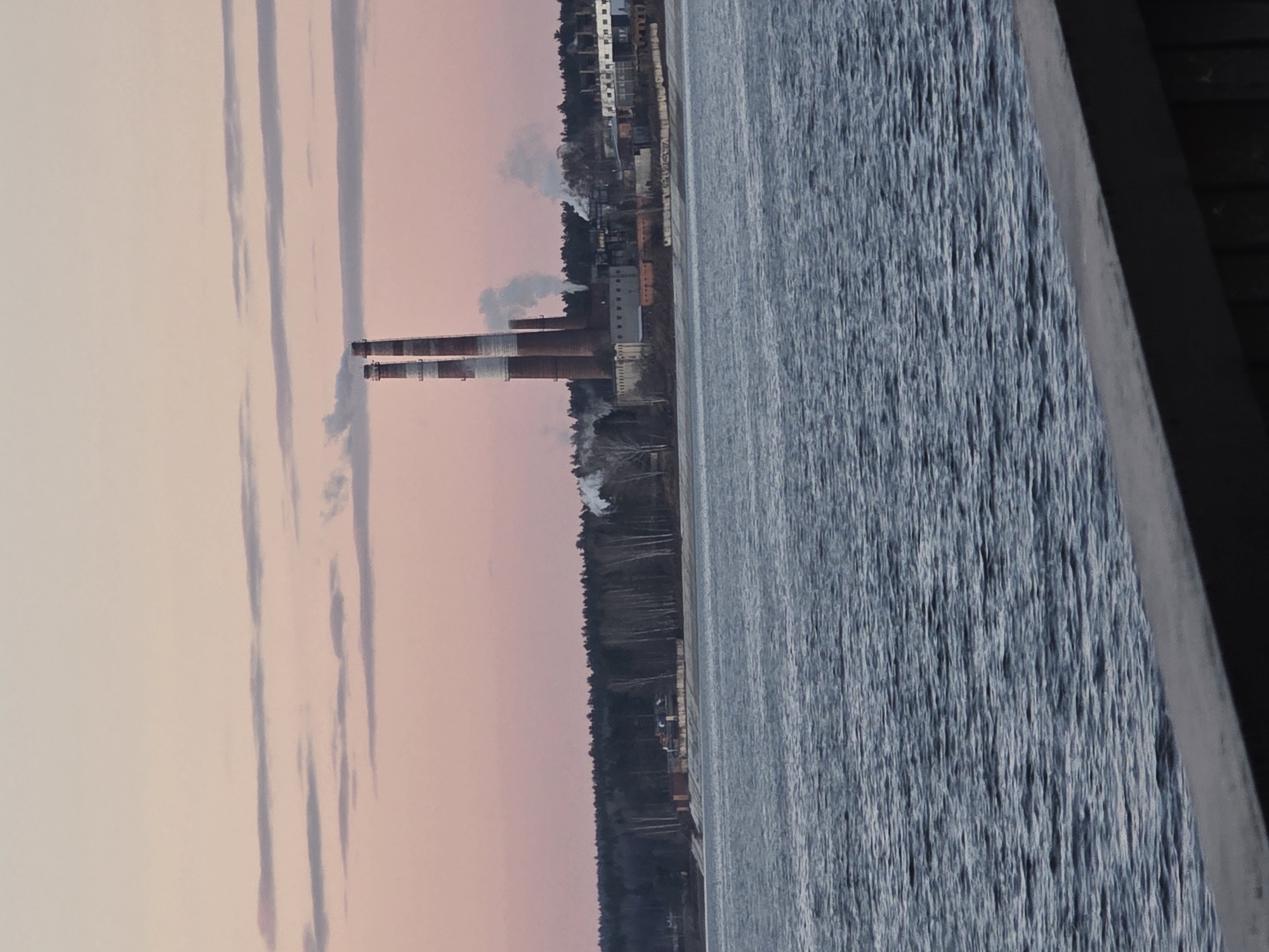 The image displays an industrial landscape viewed across a body of water, likely at twilight. Two prominent, tall industrial chimneys, striped red and white/grey, stand on the distant bank, emitting plumes of white and grey smoke that rise into the sky and drift horizontally. Behind the chimneys and extending along the bank are various industrial buildings and structures, interspersed with a dense line of trees forming a dark forest edge. To the right, a wide expanse of water, likely a river or lake, fills the foreground and midground, appearing textured with small ripples. A dark, angular structure, possibly a bridge railing or dock, occupies the extreme right edge of the frame.
The scene captures ongoing industrial activity, indicated by the smoking chimneys. The time of day appears to be late afternoon or early evening, characterized by the soft, pastel pink and light grey hues of the sky, suggesting either a serene sunset or sunrise. The smoke trails are distinctly visible against this colored sky. The water has a cool, blue-grey tone, contrasting with the warmer sky. The overall atmosphere is tranquil yet highlights human industrial presence. There is no visible text in the image. The location is contextualized as Chaikovsky, Russia.