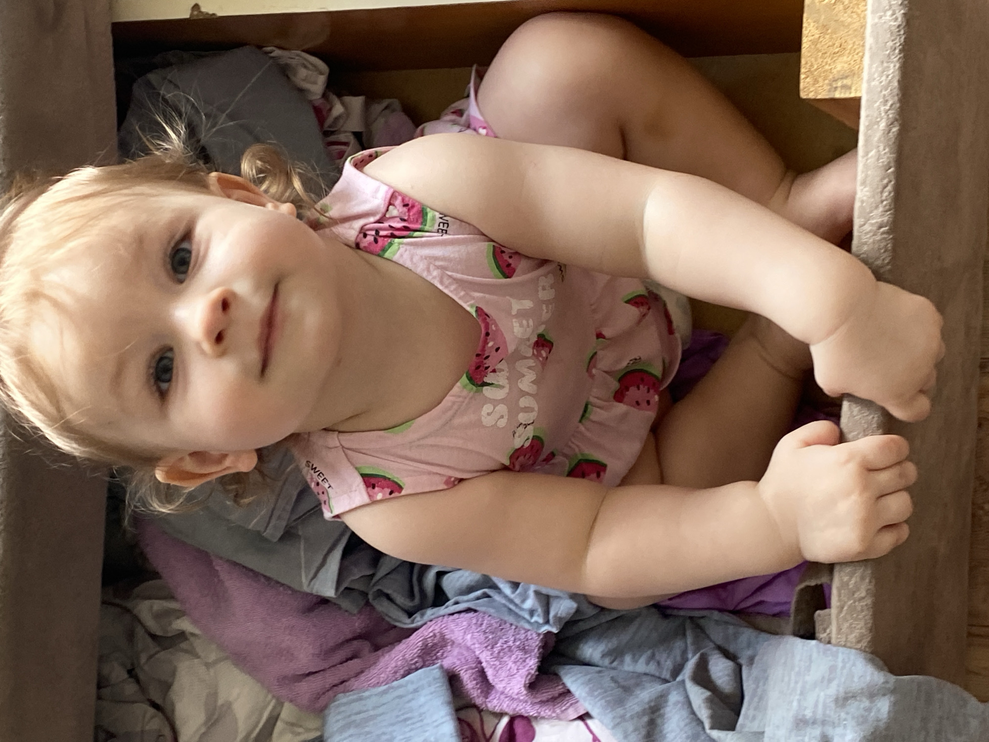 A child lies inside a wooden drawer. The child wears a pink sleeveless shirt with watermelon designs. Various articles of clothing, including gray and purple fabrics, are beneath the child. The location is Unknown, Unknown.