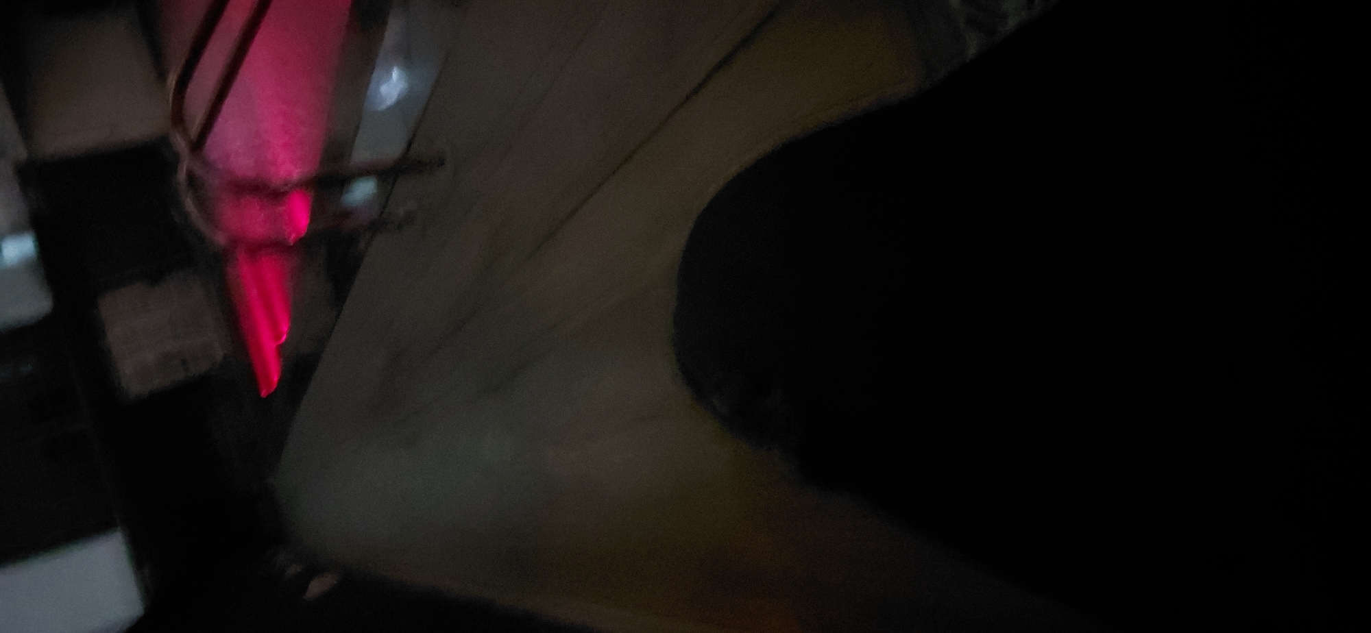 The image is a low-angle, blurred shot of what appears to be a person's leg and thigh in the foreground, covered by dark fabric. In the background, there are indistinct shapes of structures, and a bright pink light emanates from a curved object, possibly a light fixture or part of a vehicle. The scene is dark and appears to be outdoors, though the specific location in Itirin, Nigeria, cannot be determined from the provided image. The overall impression is that of an urban or semi-urban environment at night.