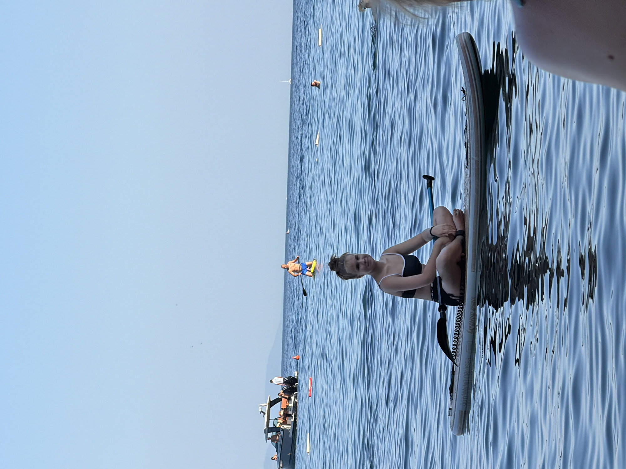 A young woman with blonde hair tied in a bun is sitting cross-legged on a paddleboard in the water. She is wearing a black bikini top and black shorts. The paddleboard has a black paddle attached to it. In the background, a boat with several people on board is visible, as well as other individuals paddleboarding and swimming. The water is calm and blue, and the sky is clear. The scene appears to be a recreational area in Budva, Montenegro.