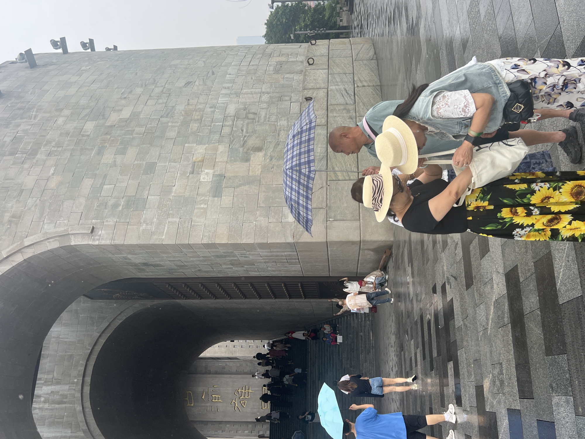 The image depicts a scene in front of a large stone structure with arched entrances in Yuzhong District, China. In the foreground, three women are interacting. One woman in a black shirt and a yellow sunflower print skirt is holding a white tote bag. Another woman in a denim vest over a white lace top and a floral print skirt is reaching into the tote bag. A third woman, wearing a straw hat with a black and white striped ribbon, is also looking at the tote bag. To their right, a man wearing a light green shirt is holding a blue and white checkered umbrella. The ground is paved with dark and gray stones, and it appears to be wet, suggesting recent rain. Further back, a blue umbrella is held by a person wearing a blue t-shirt and black shorts. A set of stone stairs leads up into one of the arched entrances, with several people walking up the stairs. The architecture of the stone structure is imposing, with large blocks and an arched opening that leads into a darker interior. The overall atmosphere is overcast, possibly due to rain.