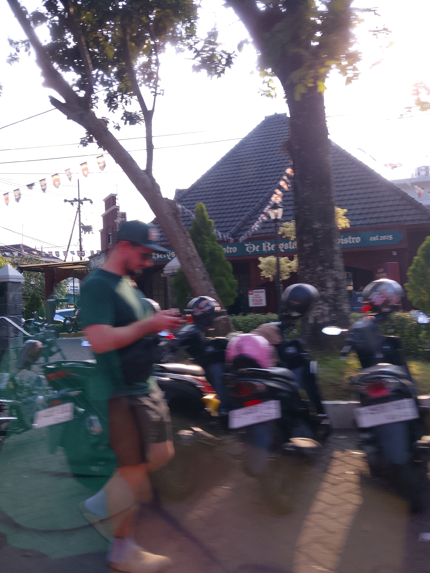 This image depicts a street scene in Malang, Indonesia, with a focus on a man walking in the foreground. He is wearing a green t-shirt, shorts, and a baseball cap, and appears to be looking at his phone. Behind him, several motorcycles are parked, and a building with a dark, tiled roof and a sign reading "De Bergsta Bistro est. 2005" is visible. The sun is setting, casting long shadows across the street, and decorative flags are strung across power lines in the background. The overall atmosphere is casual and everyday, suggesting a typical afternoon in a bustling Indonesian city.