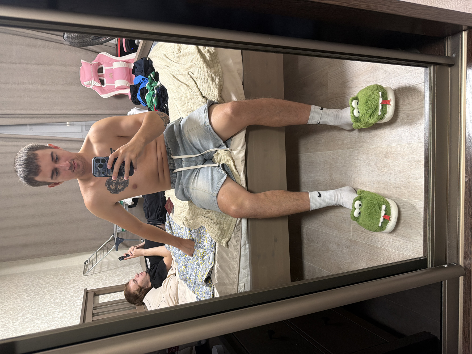 The image shows a bedroom. A young man is taking a mirror selfie. He is shirtless and wearing light blue denim shorts and white socks with black Nike logos. He has a tattoo on his chest and arm. His feet are adorned with fuzzy green alligator slippers. He is holding a mobile phone. In the background, another young man is lying on a bed, looking at his phone. He is wearing a black t-shirt and dark pants. The room has a bed with patterned bedding, a pink gaming chair, a pile of clothes, and a standing fan. The overall impression is of a casual, indoor setting. The location is not specified in the image.