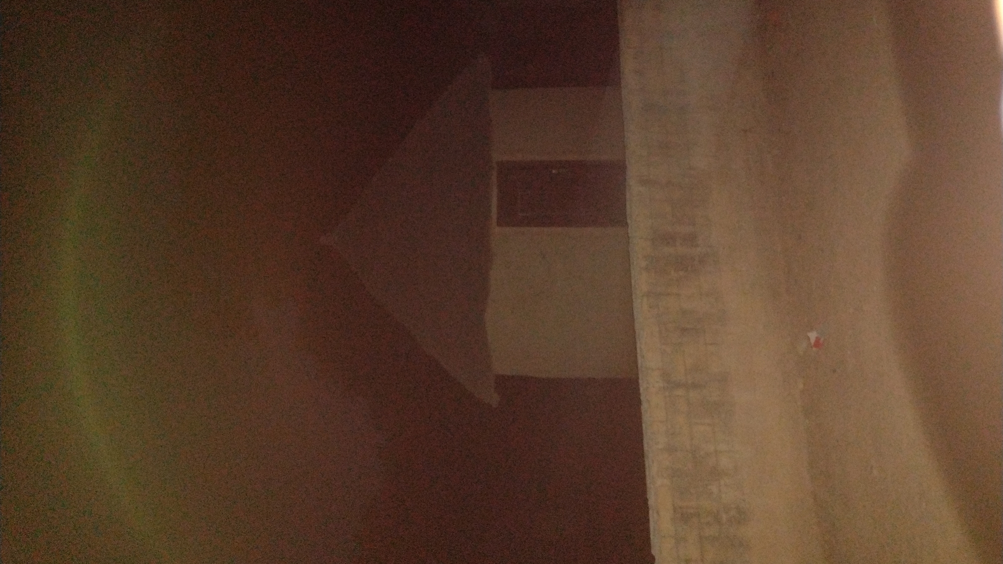 The image shows an interior view of a room in Jos, Nigeria. The foreground is obscured by shadows. A concrete structural element is visible in the right side of the frame. Adjacent to the concrete pillar, a small scrap of paper, marked in red, adheres to the wall. Beyond the shadowed foreground, a wall section appears with a dark rectangular feature at the top. The composition suggests a dimly lit room.