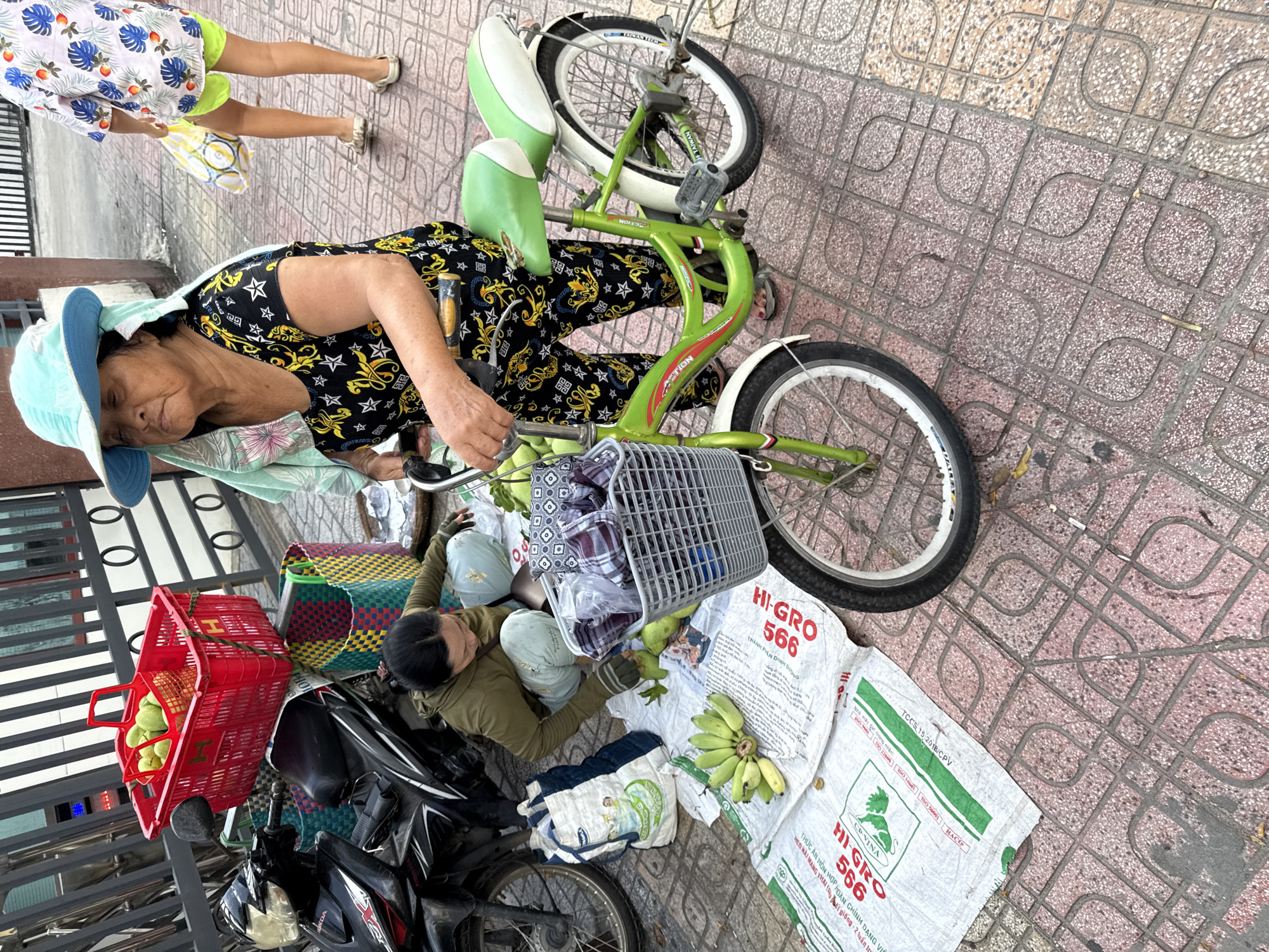 In Nha Trang, Vietnam, an elderly woman wearing a light blue sun hat and a black and gold patterned shirt is sitting on a green and white bicycle on a sidewalk paved with red and beige tiles. She is holding the handlebars of the bicycle, which has a basket on the front containing checkered fabric. To her left, another woman in a green jacket is kneeling on the ground, arranging produce. A black motorcycle is parked to the left of the women, with a red basket filled with green fruit on its seat. Several woven bags are scattered on the ground. The scene appears to be a street market, with various items laid out for sale.