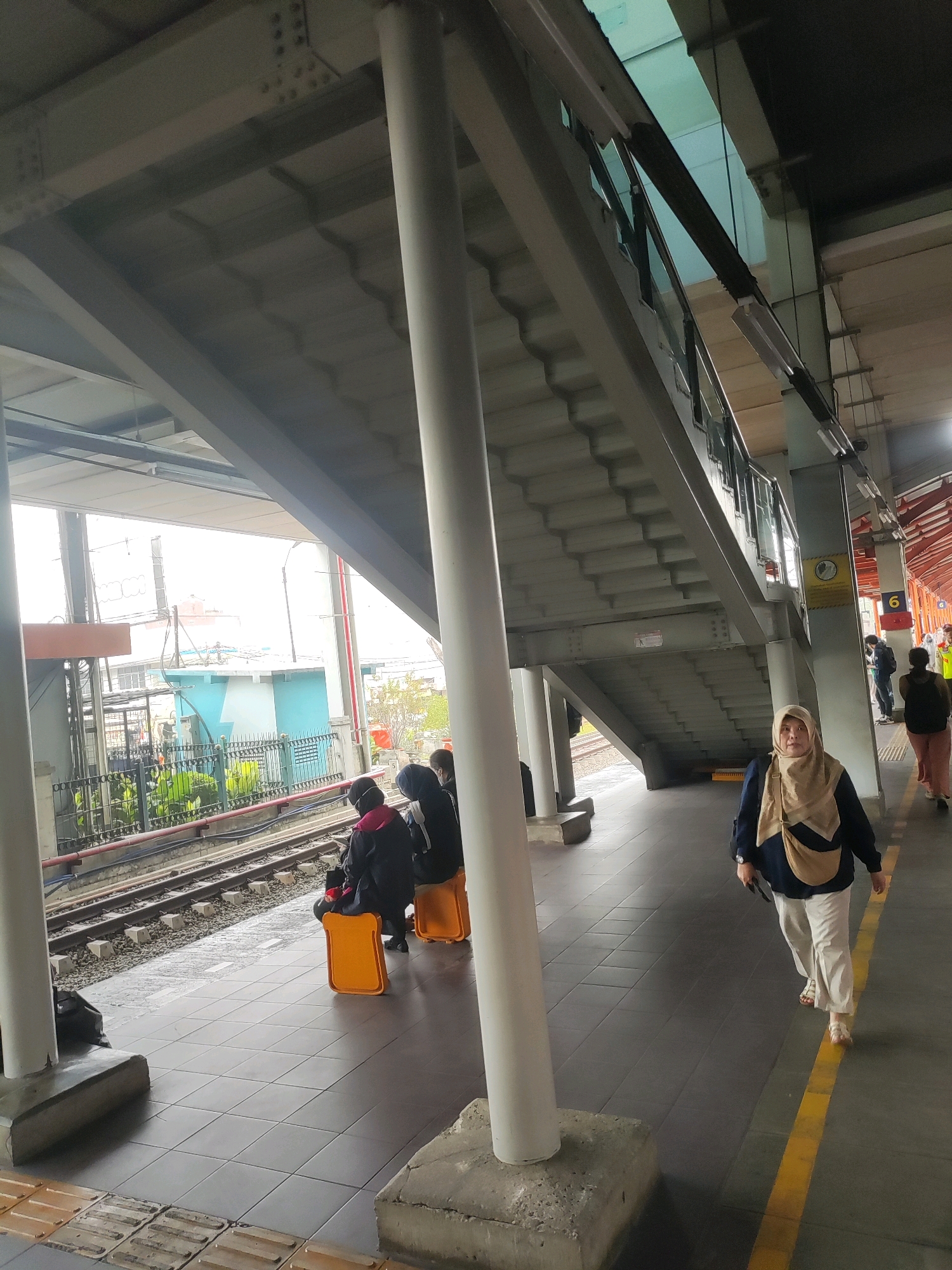 The image depicts an outdoor platform at a train station in Bekasi, Indonesia, with a pedestrian overpass structure dominating the scene. The overpass, made of concrete and steel, features a wide staircase ascending to the right. Below the overpass, a train track runs horizontally across the frame.
On the platform, several individuals are visible. In the foreground, a woman wearing a beige hijab and cream-colored pants is walking towards the right, her gaze directed slightly upwards. She carries a bag slung across her shoulder. Further back, a group of three people are seated on bright orange, bread-shaped stools near the train tracks, facing away from the camera. They appear to be waiting.
Beyond the tracks, a building with teal and white accents is visible, surrounded by green foliage behind a dark metal fence. In the distance, other structures and overhead electrical wires suggest an urban environment. The platform itself is paved with square tiles, and a yellow line marks the edge. Various support pillars for the overpass are scattered throughout the scene. Other individuals can be seen in the background, one of whom is wearing a bright yellow vest, potentially an employee. The overall lighting suggests it is daytime, with diffused light casting shadows.