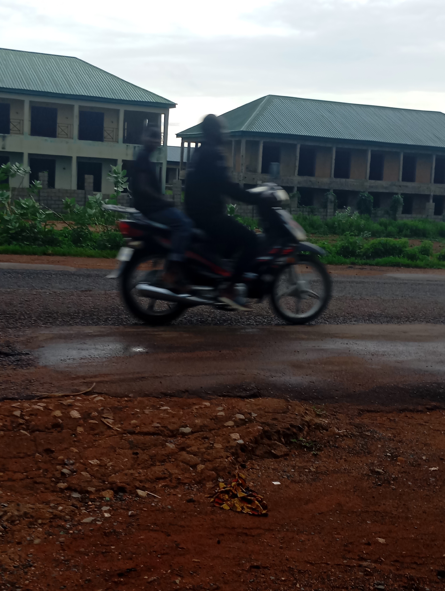 The image captures a street scene in Shendam, Nigeria, featuring two individuals riding a motorcycle. The motorcycle is in motion, causing motion blur on the subjects and the vehicle. The road surface appears to be asphalt with visible water puddles, indicating recent rain. In the background, there are unfinished buildings with green corrugated metal roofs, suggesting construction is ongoing or halted. Lush green vegetation surrounds the buildings and lines the side of the road. The sky is overcast with a cloudy sky. The foreground shows a dirt path with scattered stones and some dry leaves.