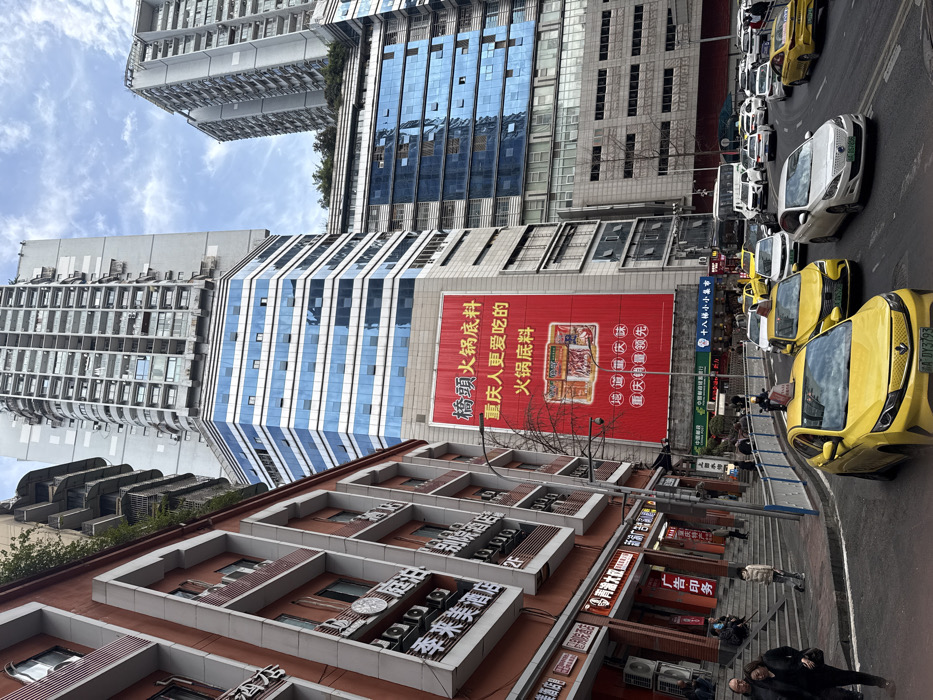 The image shows a street scene in China with tall buildings and traffic. There is a red advertising banner for “Qiao Tou Huo Guo Di Liao”, promoting Chongqing hot pot. Several yellow and white vehicles are on the road, and people are walking on the sidewalk. The buildings have a mix of architectural styles, with some featuring mirrored windows and others with traditional Chinese designs. Several businesses are visible including a hotel and printing company. The text on the advertising banner is in Chinese.