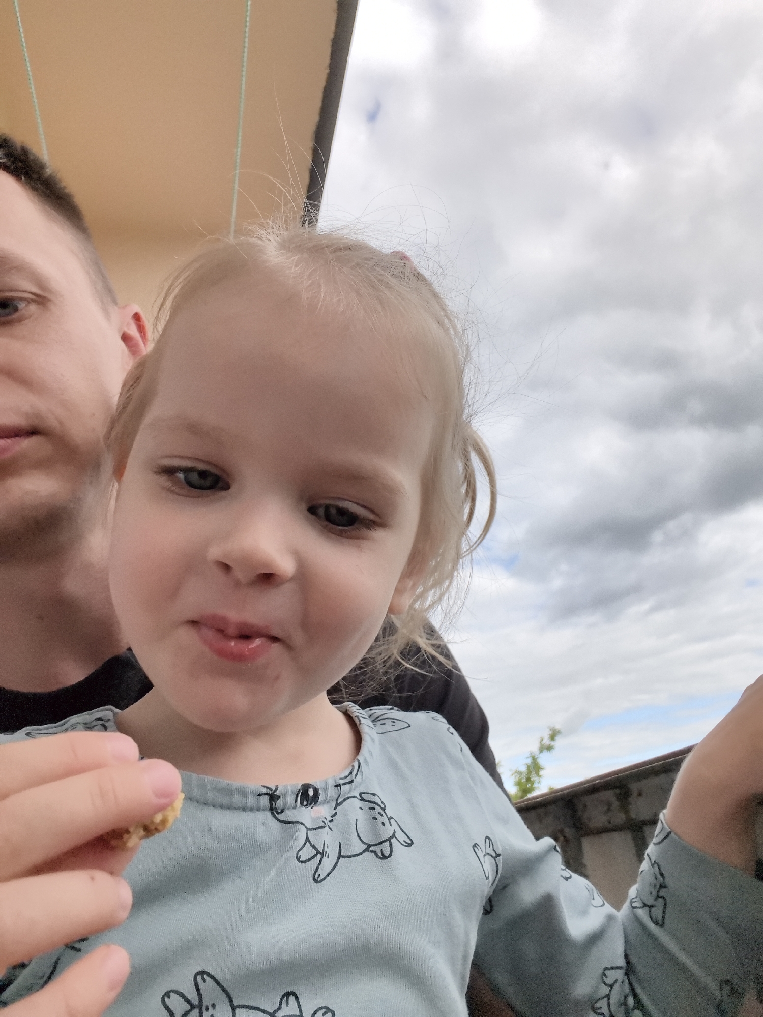A man and a young child are positioned in the foreground of the image, with the child slightly closer to the camera. The man's left side of his face, including his eye and ear, is visible. The child, facing the camera, has light-colored hair and is wearing a light blue long-sleeved shirt with a cartoon animal pattern. The child is holding a small piece of food in their right hand, near their mouth. The background consists of a cloudy sky and a blurred suggestion of an outdoor environment with some greenery. The lighting suggests it is daytime.