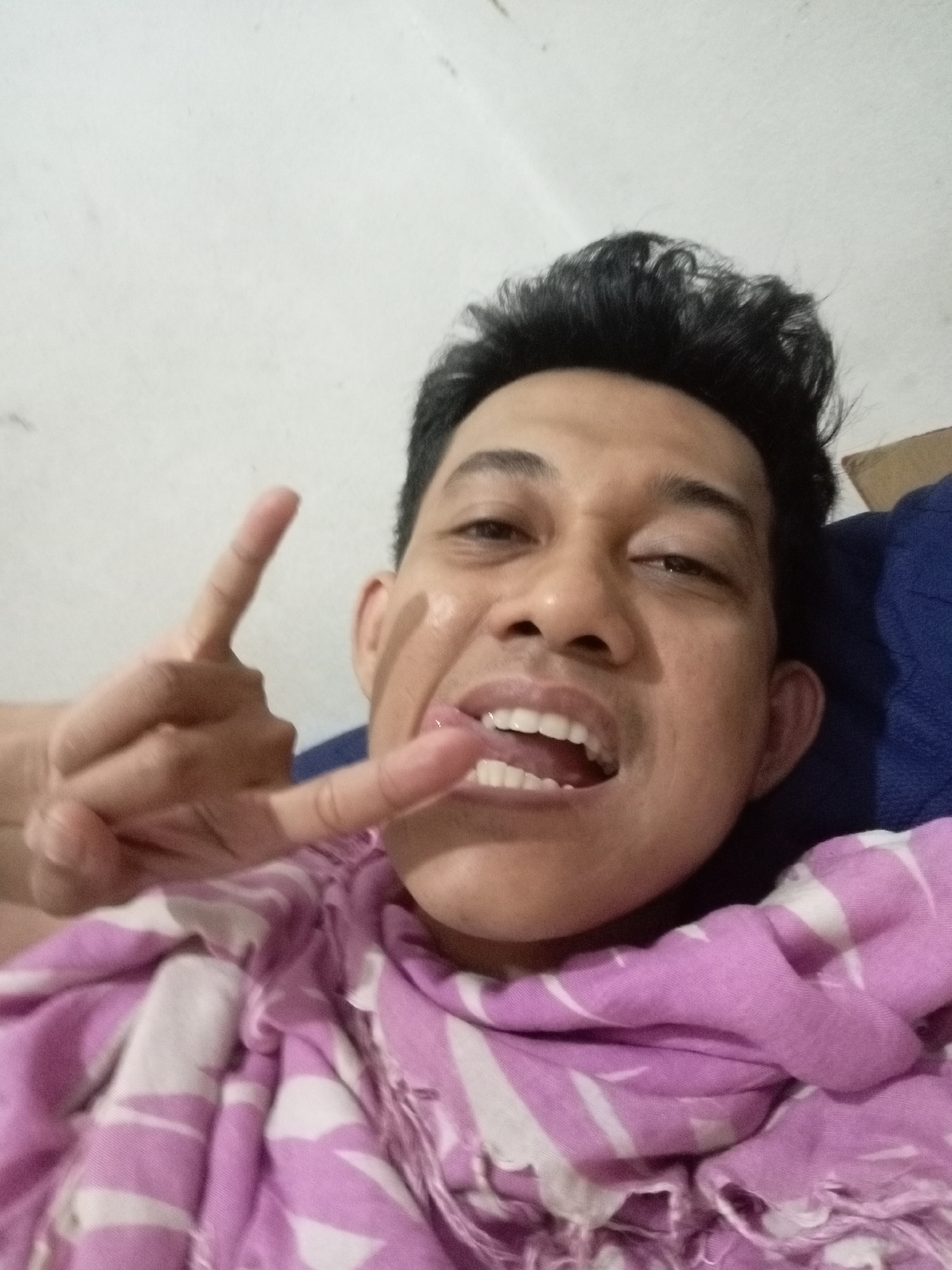 A selfie taken by a man with dark, wavy hair and light brown skin. He is wearing a pink and white striped garment. His right hand is raised, with two fingers extended in a "rock on" gesture, while his tongue is sticking out of his mouth. The background consists of a white wall. The image appears to be taken indoors, likely in a residential setting in Batam, Indonesia.