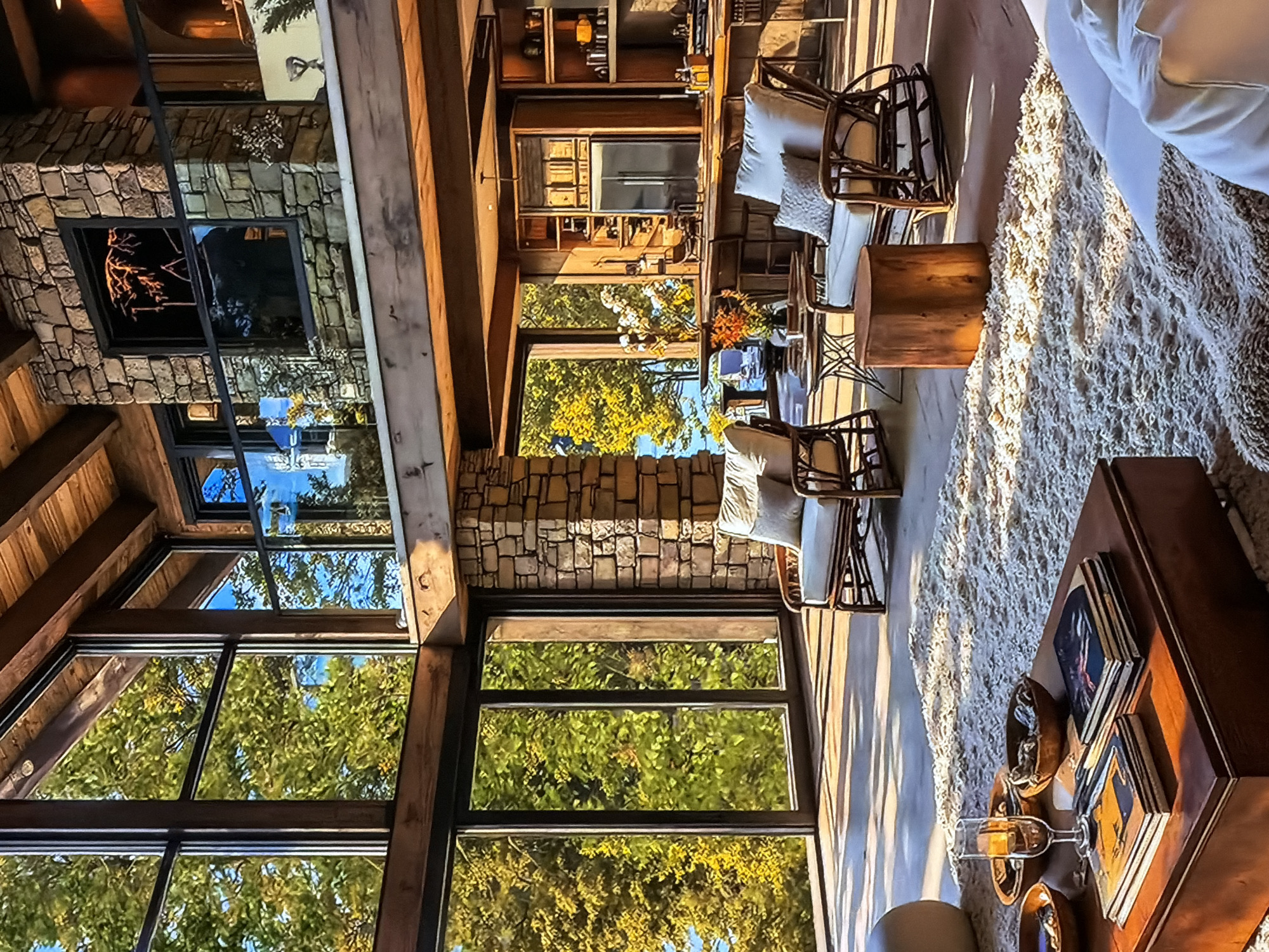 This image captures the interior of a luxurious living room, showcasing a blend of rustic and modern design elements in what is likely a high-end residence in Sofiivska Borshchahivka, Ukraine. The room is brightly illuminated by abundant natural light streaming through expansive, multi-pane windows that span from floor to ceiling, offering lush views of green trees and foliage, suggesting a serene natural setting.
Architectural features include prominent wooden beams on the ceiling and as structural supports, contrasting with stone-clad walls, particularly around a large, modern fireplace on the left side of the frame. The fireplace features a dark, reflective insert.
The living area is furnished with several rattan or woven chairs, each adorned with plush, light-colored cushions. These are arranged around low wooden tables, one of which holds a vase of vibrant orange and yellow flowers. A large, shaggy, light-toned area rug covers a significant portion of the floor, adding texture and warmth to the space. In the foreground, another dark wooden coffee table displays stacked books, decorative bowls, and a clear wine glass, indicating a space set up for relaxation. In the background, a substantial wooden cabinet or shelving unit appears to be a bar or display area.
The strong, clear sunlight casting shadows across the rug suggests it is a bright, sunny day, likely late morning or early afternoon. No people are visible, and no specific activity is taking place, but the arrangement of furniture implies a comfortable space for socializing or unwinding. The overall atmosphere conveys tranquility, comfort, and a strong connection to the surrounding nature. There is no visible text in the image.