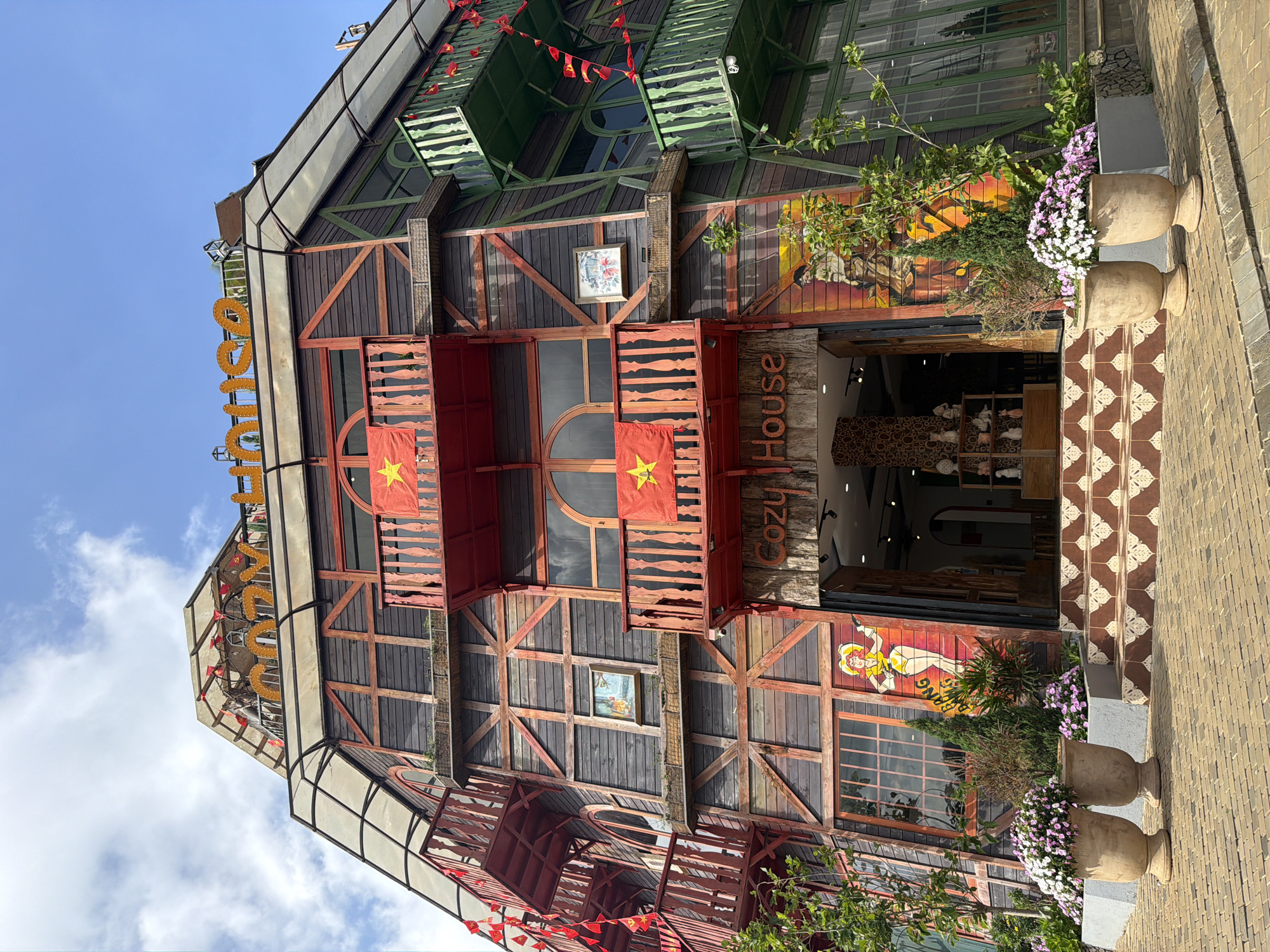 This image captures the vibrant, multi-story facade of a building, rotated 90 degrees clockwise from its upright orientation, under a clear, sunny sky. The structure features a distinctive architectural style, combining dark wood paneling with red wooden cross-beams and green slatted sections, possibly balconies or decorative roof elements.
Large golden letters spelling "MANG DEN" are prominently displayed along the roofline, with strings of red pennant flags draped below. Multiple balconies with red railings are visible, each adorned with the national flag of Vietnam (red with a yellow star). The building's windows vary in shape, some rectangular, others arched, integrated into the wooden framework.
To the right of the main entrance, which offers a glimpse into a dimly lit interior with shelves displaying various items, a vertical sign spells "Cozy House." Below this, a colorful mural depicting a woman in a yellow dress adds an artistic touch to the facade. Further decorative elements include small framed pictures on the wooden walls.
At the ground level, a patterned walkway of brown and white tiles leads to a brick-paved path. Two large, light-colored planters are filled with a lush display of white and purple flowers. The bright, clear sky indicates a sunny daytime setting. The scene suggests a unique cafe, shop, or guesthouse, characteristic of its Măng Đen, Vietnam location.