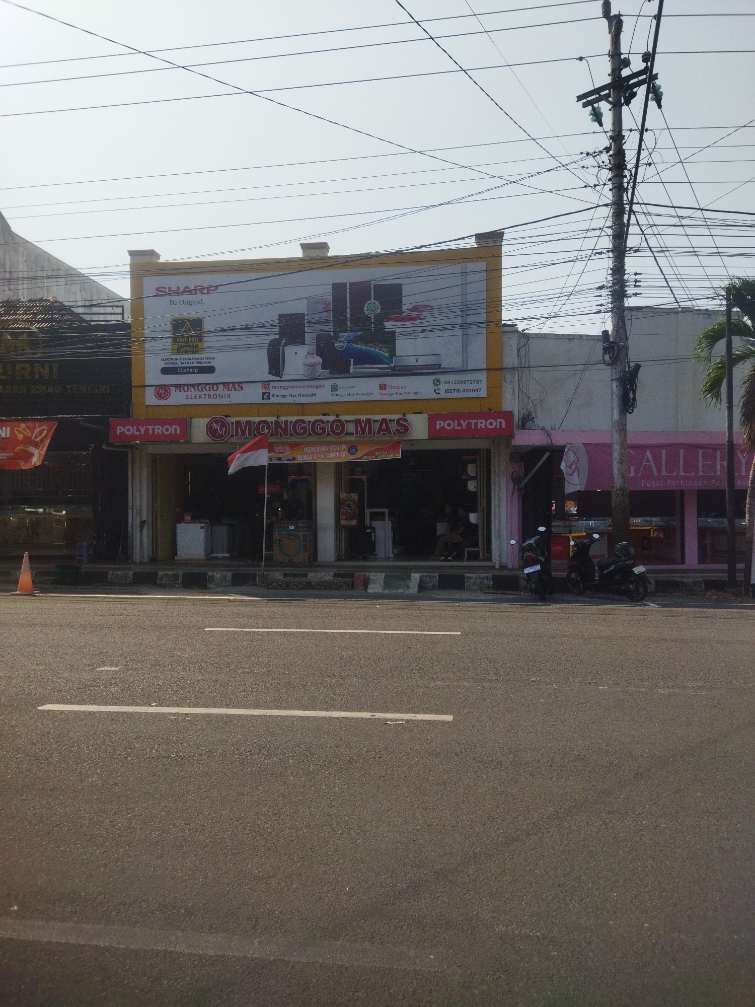The image depicts a street scene in the city of Wonogiri, Indonesia. In the foreground, a paved road with white lane markings stretches across the frame. A sidewalk with a curb separates the road from the buildings. On the sidewalk, an orange traffic cone is visible.
The main focus of the image is a row of commercial buildings. The most prominent is a store with a large yellow and white banner displaying "SHARP Be Original." Below this banner, the store name "MONGGO MAS ELEKTRONIK" is displayed in large red and white letters. The store's facade features multiple storefronts, with the central one displaying a red "POLYTRON" sign. A small Indonesian flag is positioned in front of the "MONGGO MAS" storefront. Two motorcycles are parked on the sidewalk in front of the buildings.
To the right of "MONGGO MAS ELEKTRONIK," another storefront with a pink awning displays the word "GALLERY" in large lettering, with the tagline "Pusat Perhiasan Peraga" below it, indicating it is a jewelry store. A palm tree is visible to the far right of the buildings.
The sky is clear and bright, suggesting a sunny day. Overhead, a complex network of electrical wires and a utility pole are prominent features of the urban environment. The overall impression is a typical street scene in an Indonesian city, with commercial activity and infrastructure.