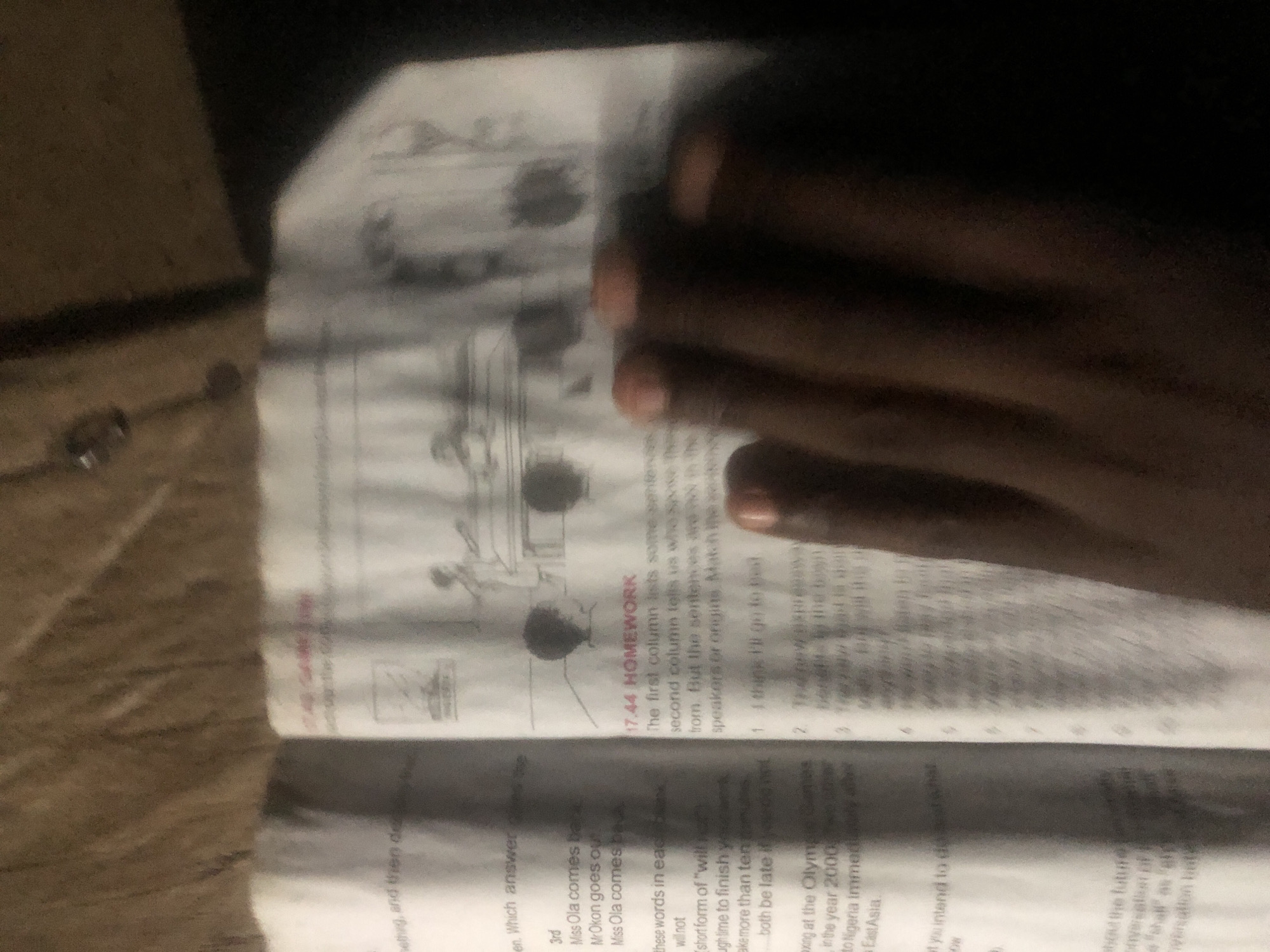 The image shows a close-up, blurry view of an open book, likely a textbook or workbook, placed on a textured, brown surface. The book is open to a page with text and illustrations. On the left page, there are several lines of text, some of which appear to be questions or exercises. One section mentions "Miss Ola" and "Mr Okon." Further down, there's a paragraph about "boxing at the Olympic Games" in the year 2000, and mentions Nigeria. The right page contains a heading "17.44 HOMEWORK" followed by instructions and a list of numbered sentences. There are also some simple line drawings of people and furniture at the top of the right page. A person's dark hand, with visible fingers and fingernails, is resting on the right page of the book, obscuring parts of the text and illustrations. The lighting appears to be dim, contributing to the blurriness of the image. There is no discernible information about the city of Jalingo, Nigeria, within the visible content of the image.
