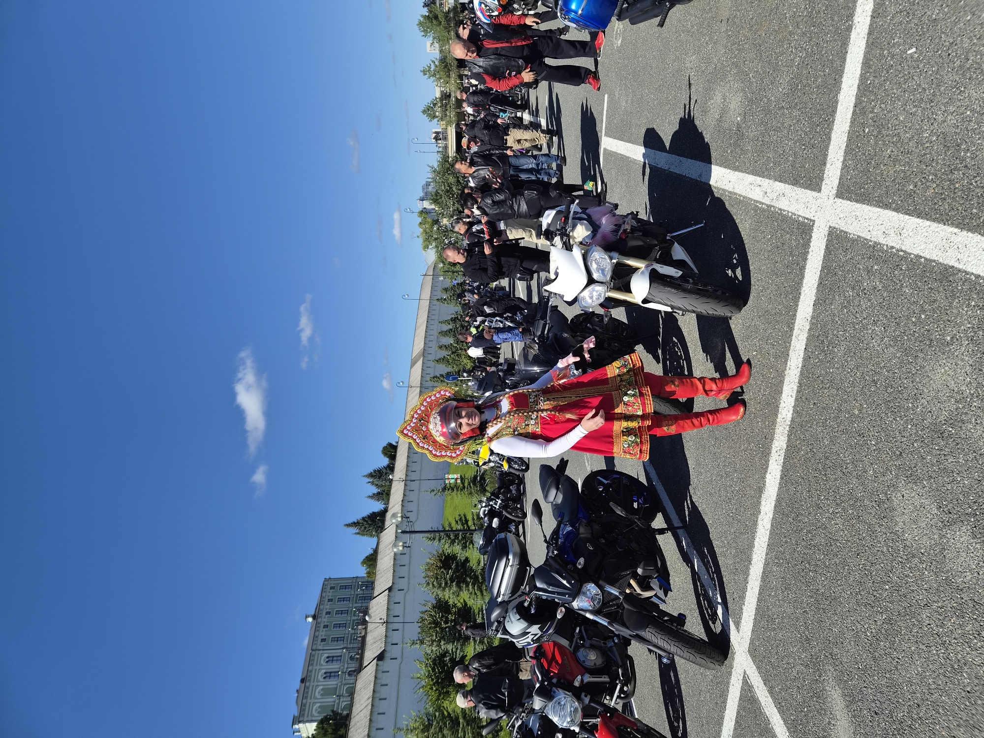 In Kazan, Russia, under a clear blue sky with scattered clouds, a woman dressed in a traditional red and gold Russian costume stands in a parking lot filled with motorcycles. She wears a tall, ornate headdress and matching red boots. Several motorcycles, including red, blue, and white models, are parked around her. In the background, a long, white fortified wall with arched windows stretches across the landscape, with trees and buildings visible beyond it. A crowd of people, many wearing leather jackets, are gathered near the motorcycles, some engaged in conversation or looking at their phones. The scene appears to be an outdoor event, possibly a motorcycle rally or festival, taking place in Kazan.