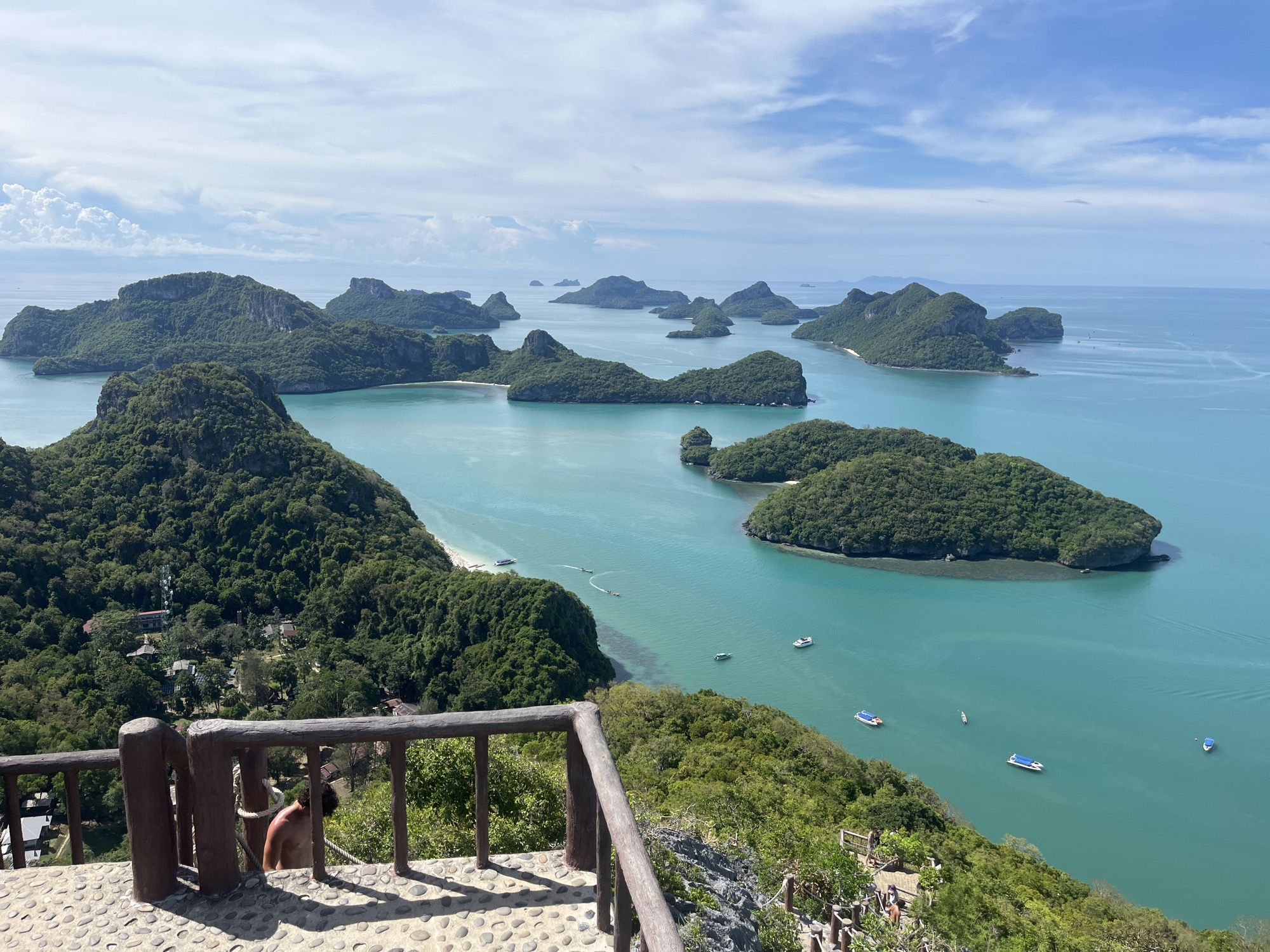 This is a high-angle, wide shot of a tropical archipelago on a bright, partly cloudy day. The foreground features a weathered wooden railing and a stone pathway leading down a steep, vegetated slope. A person with tanned skin and curly hair, wearing only shorts, is visible leaning against a rope by the railing. Further down the path, a few other people are seen walking.
The midground and background are dominated by numerous verdant, mountainous islands rising from turquoise-blue waters. Several small boats, including speedboats and a longtail boat, are scattered across the sea. Some islands have small white-sand beaches. The water is clear, revealing shallow areas near the shorelines. In the distance, more islands are visible, stretching towards the horizon under a bright blue sky with scattered white clouds. The scene is likely a viewpoint in Ang Thong National Marine Park, Koh Samui, Thailand, known for its dramatic limestone formations and clear waters.