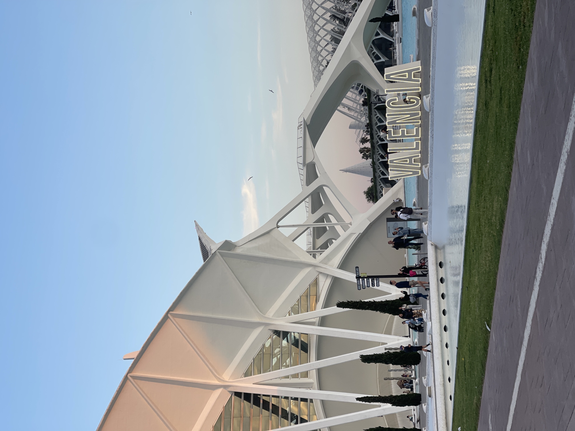 The image depicts the City of Arts and Sciences complex in Valencia, Spain. The foreground includes a prominent white architectural structure with geometric lines and a triangular roof. A large "VALENCIA" sign is positioned near a shallow pool of water. People are visible along a walkway next to the pool. In the background, another building with a lattice-like structure can be seen. The sky is blue with a few birds flying.