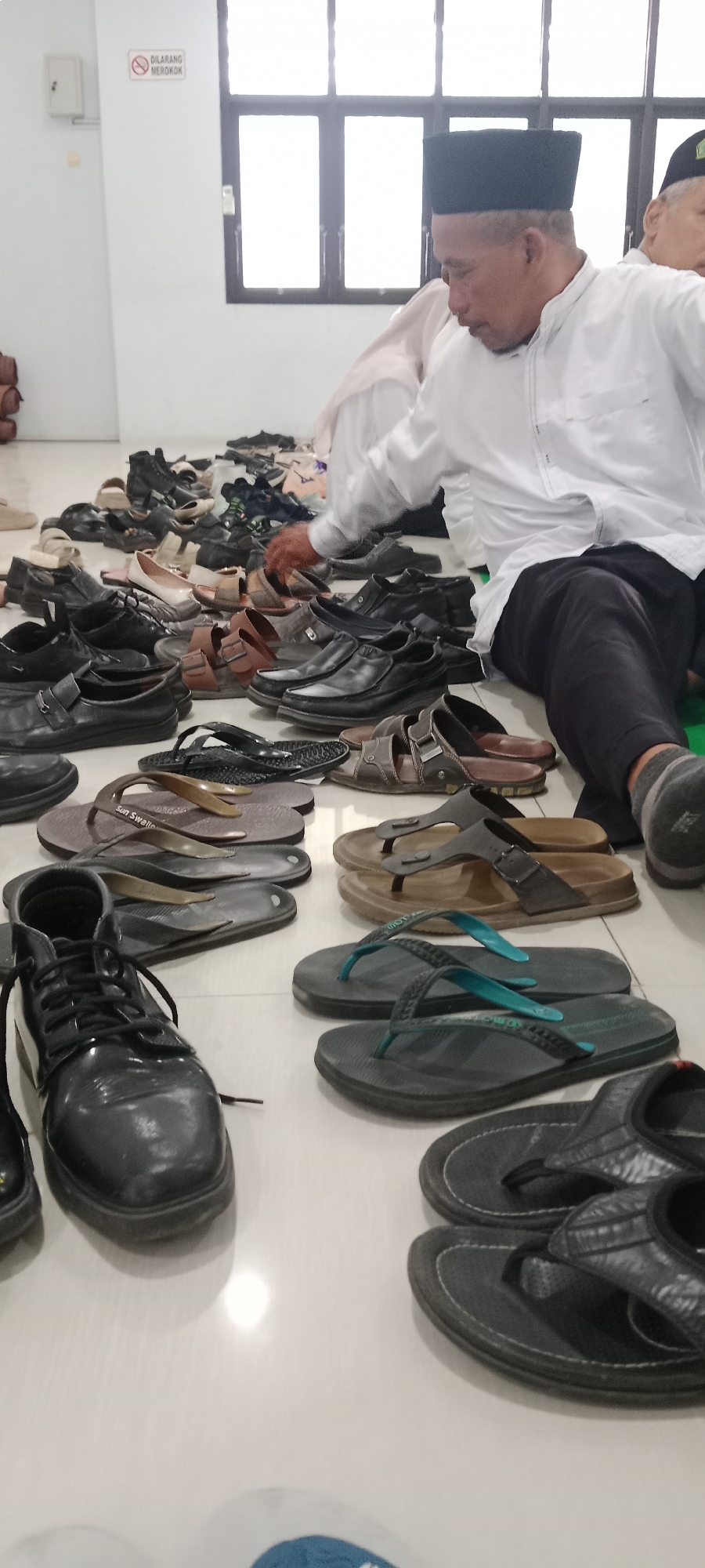 The image depicts an indoor scene, likely in the city of Tuban, Indonesia, with a significant collection of footwear arranged on a light-colored tiled floor. In the foreground, a black leather boot with laces is prominently displayed. Various pairs of sandals and flip-flops are scattered throughout the scene, in shades of brown, black, and grey, with some featuring colorful straps. Several pairs of men's dress shoes, primarily black, are also visible.
In the background, a man wearing a white long-sleeved shirt, black pants, and a black peci (a type of cap) is seated on the floor. He is looking down, and his right hand is extended, appearing to interact with some of the shoes. Another person, partially visible to the right, also appears to be wearing dark pants and a grey sock. The environment includes a plain white wall and a window with multiple panes. A small white sign with red lettering and a prohibition symbol is affixed to the wall, indicating "DILARANG MEROKOK" (Smoking Forbidden). The overall impression is one of a communal area where shoes are stored or gathered, possibly at a place of worship or a public facility in Tuban.
