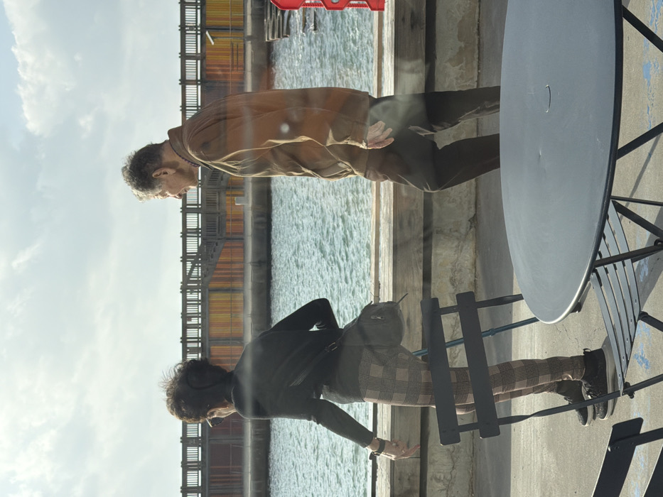 The image depicts an outdoor scene at the Tel Aviv Port (Namal Tel Aviv) in Tel-Aviv, Israel, under bright, partly cloudy daylight. Two elderly individuals are walking from right to left across a concrete promenade adjacent to the Mediterranean Sea.
In the immediate foreground, dark metal outdoor cafe furniture is visible, including a partially cropped round table on the right and a dark metal chair on the left. A blue marking is on the concrete ground near the chair.
The individual on the left is a female, seen from behind and slightly to her right. She has short, dark, curly hair, wears sunglasses, a black long-sleeved top, plaid leggings in brown and tan tones, and black sneakers. A black shoulder bag is slung across her body. Her left wrist bears a dark-colored watch, and her right hand is partially extended with fingers slightly curled. A blue/black walking cane is partially visible, appearing to lean against the foreground chair.
The individual on the right is a male, seen in profile, walking slightly ahead of or alongside the female. He has short, grey, curly hair and wears a brown long-sleeved jacket and dark trousers. His right arm is bent at the elbow.
Between the promenade and the background structure, a body of water with moderate ripples is visible. A low wooden barrier runs along the edge of the promenade. The background is dominated by the distinctive wooden deck and support structure of the Tel Aviv Port. Below the wooden deck, a wall is decorated with vertical panels in shades of orange, yellow, red, and brown. A wooden staircase ascends onto the pier structure on the left side of the background. Sun glare or reflections are present across various parts of the image, suggesting it may have been photographed through glass.