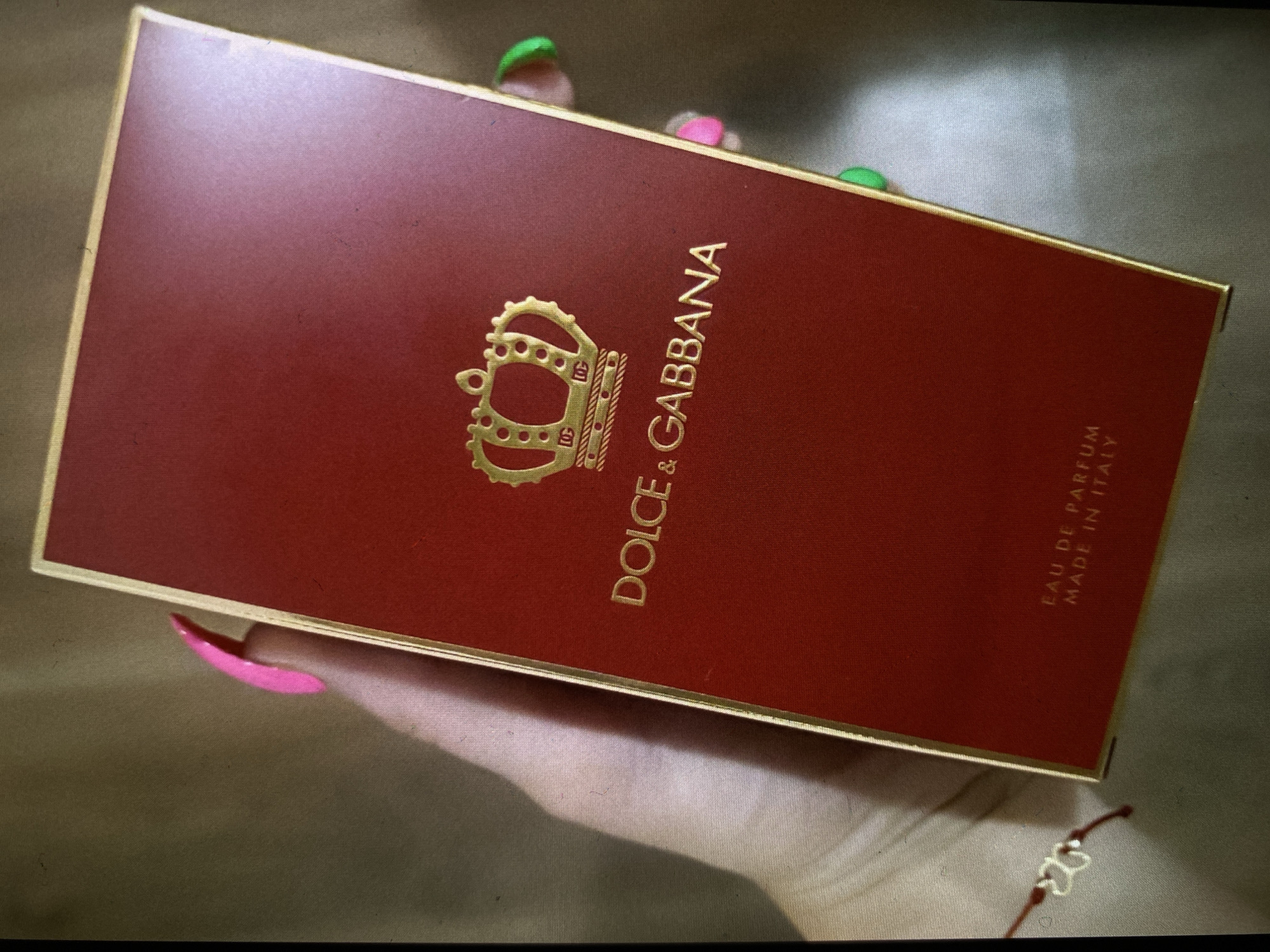 A close-up, slightly angled shot shows a person's hand holding a rectangular, dark red box, likely packaging for a luxury product. The box features a prominent gold crown logo on its left side. To the right of the crown, the brand name "DOLCE & GABBANA" is printed vertically in gold capital letters. Further to the right, also in gold and vertical, are the words "EAU DE PARFUM" and "MADE IN ITALY". The box has a subtle texture and a thin gold border around its edges.
The hand holding the box displays brightly colored nail polish: a vivid pink on the thumb and index finger, and a neon green or yellow-green on the visible tips of other fingers. A delicate red string bracelet with a small, metallic, heart-shaped charm is visible on the wrist. The background is out of focus, appearing as a mottled surface of muted brown and grey tones, suggesting an indoor setting. There's a slight glare on the upper left corner of the box, indicating a light source. The image captures the act of presenting or holding this branded item. No specific time of day, weather conditions, or direct location cues for Bankya, Bulgaria are discernible from the visual information alone.