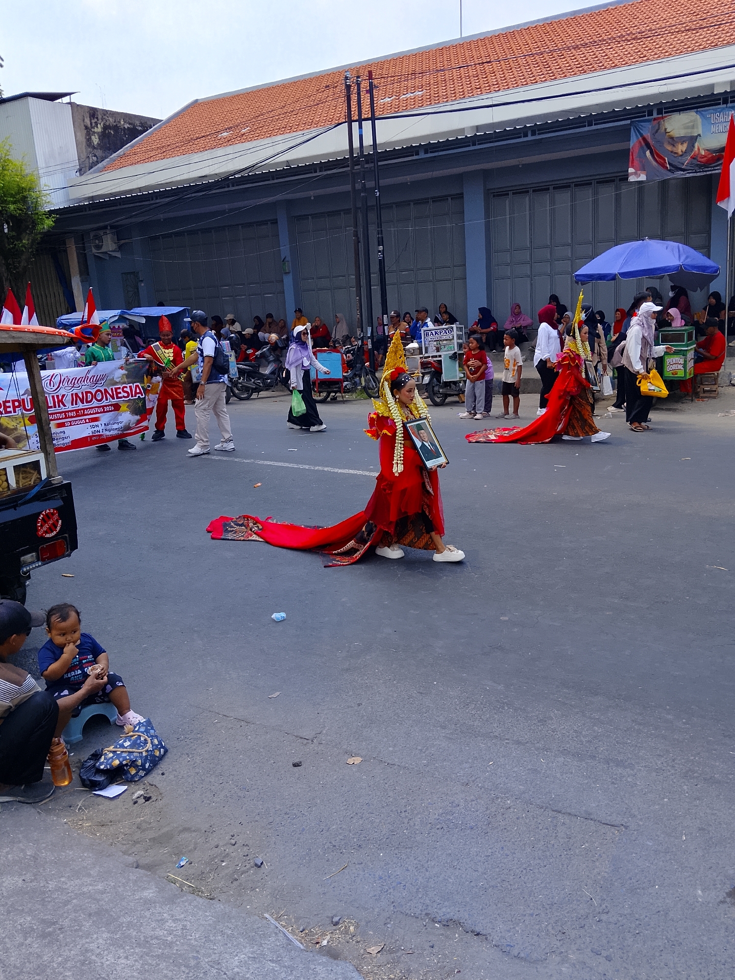 In Kertosono, Indonesia, a parade is taking place on a street lined with buildings and a crowd of spectators. Two individuals in elaborate red and gold traditional attire are the central figures, with one holding a framed portrait. In the foreground, a baby sits on a blue stool, eating, with an adult next to them. The street is paved and appears to be busy with participants and onlookers. The buildings in the background are typical of Indonesian architecture, with tiled roofs.