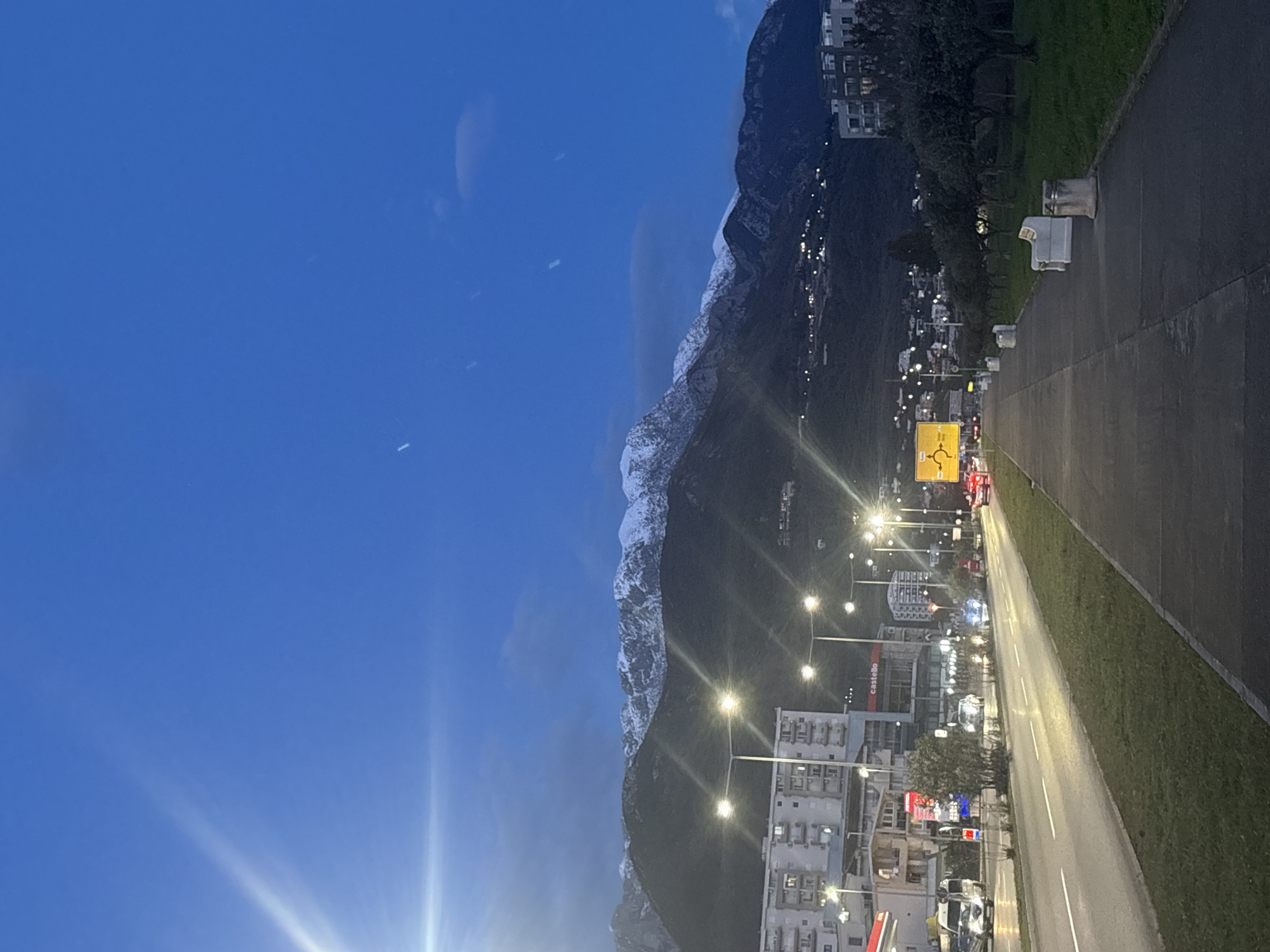 The image captures a twilight scene in Bar, Montenegro. On the right side of the frame, there's a road bordered by a grassy area. Streetlights illuminate the road, and commercial buildings line the street, some displaying signage.
In the distance, a mountain range rises, its peaks partially covered in snow. The sky transitions from a deep blue at the top to a lighter hue near the horizon. A few scattered clouds are visible. The image appears to be taken at ground level, oriented sideways.
