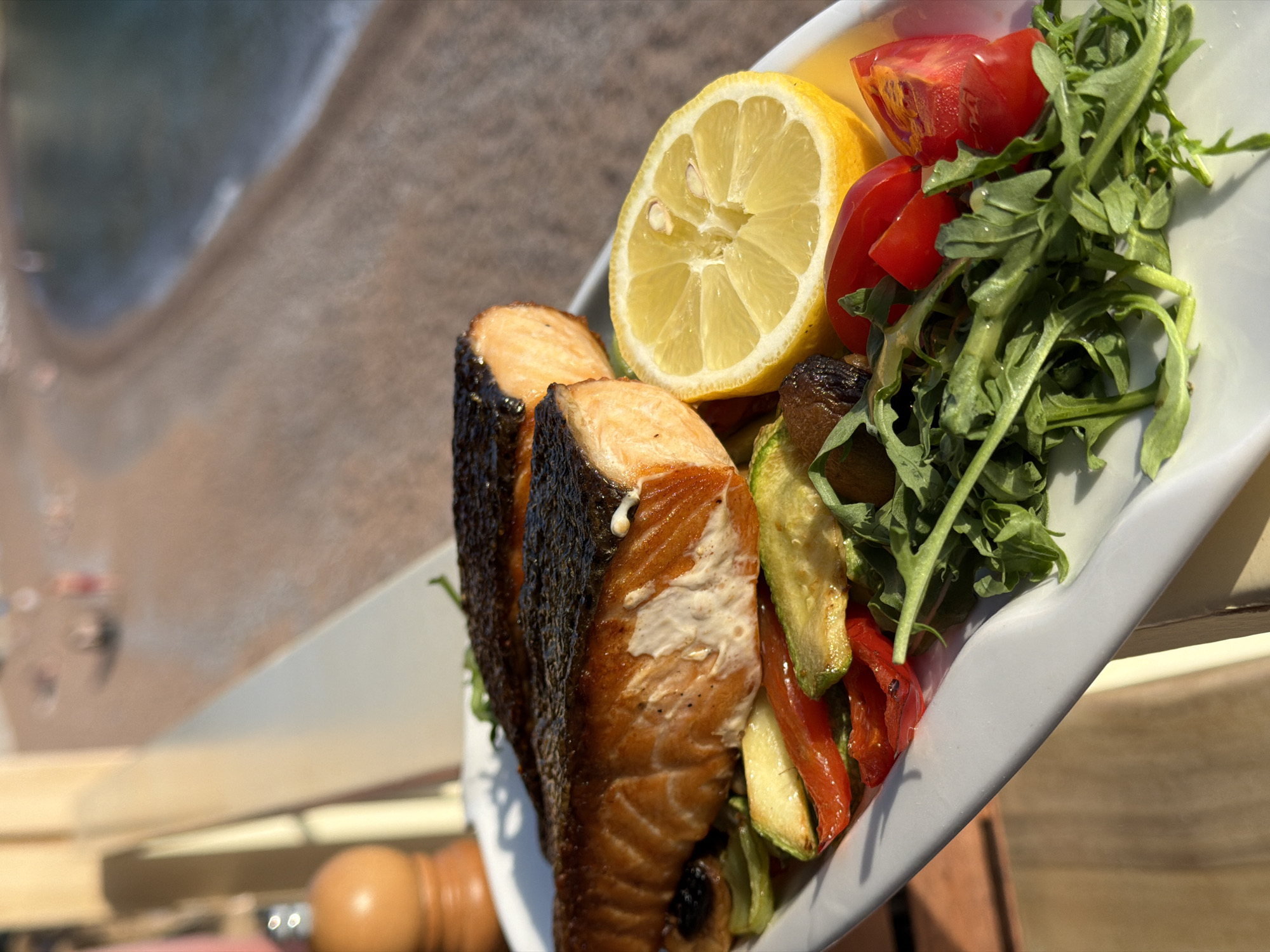 A plate of food is presented with a view of Petrovac centar, Montenegro in the background. The plate contains two pieces of cooked salmon with dark, crispy skin, a half lemon, sliced cherry tomatoes, sauteed zucchini and red bell peppers, and fresh arugula. The plate is white and appears to be held or placed on a wooden surface. The background shows a beach with people and the sea, blurred due to the focus being on the food.