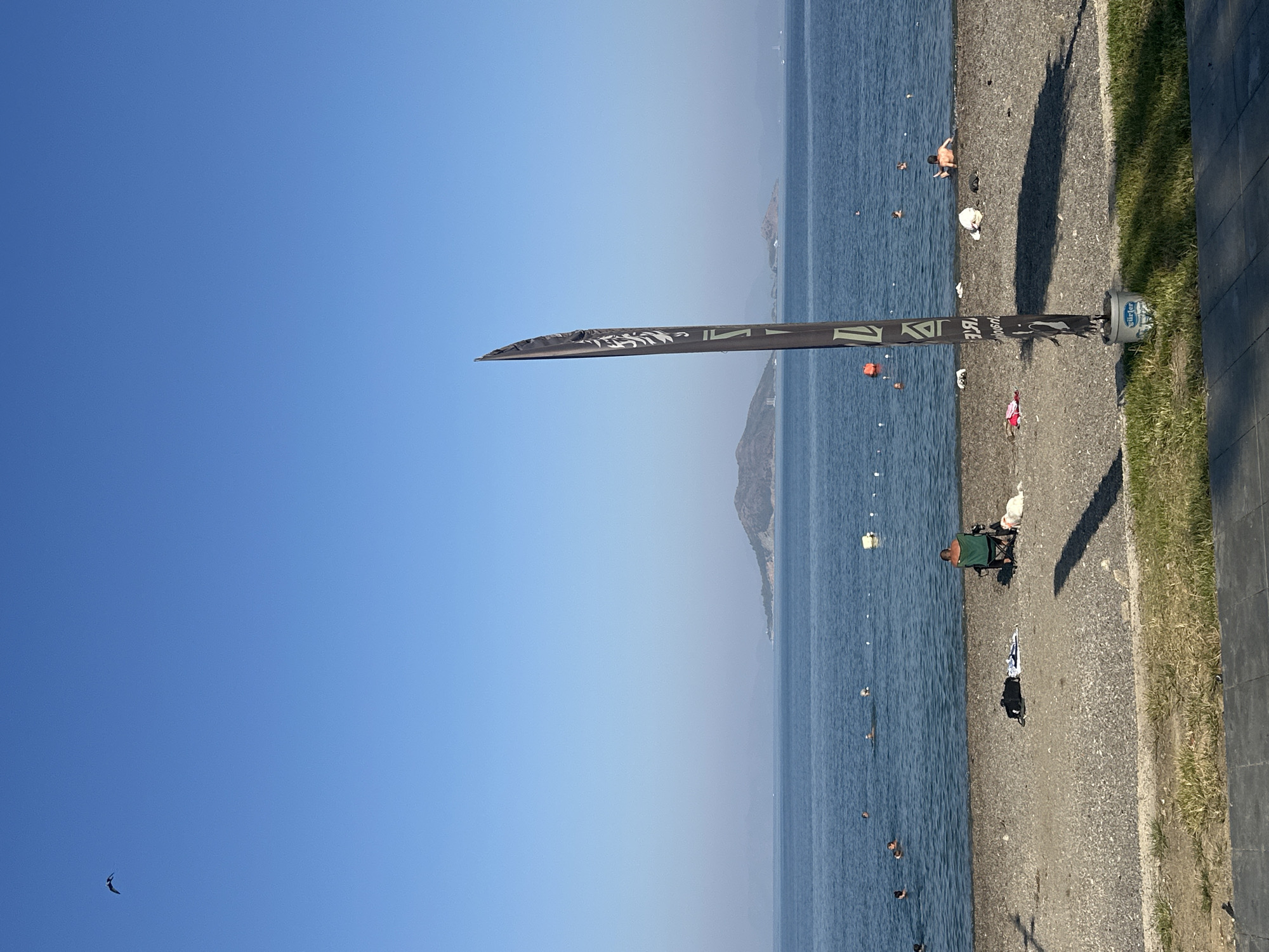 A broad view of a pebble beach on the coast of Fethiye, Turkey, under a clear blue sky. In the foreground, a tall, dark flag with white text stands on a grey bucket. To the right of the flag, the shadow of an unseen object stretches across the beach. Several beach items, including bags and towels, are scattered on the pebbles. A person sits in a green chair on the beach, facing the water, with a white dog beside them. In the middle ground, the calm blue sea is populated by a few people swimming. In the distance, a hilly island is visible on the horizon, with other land masses further away.