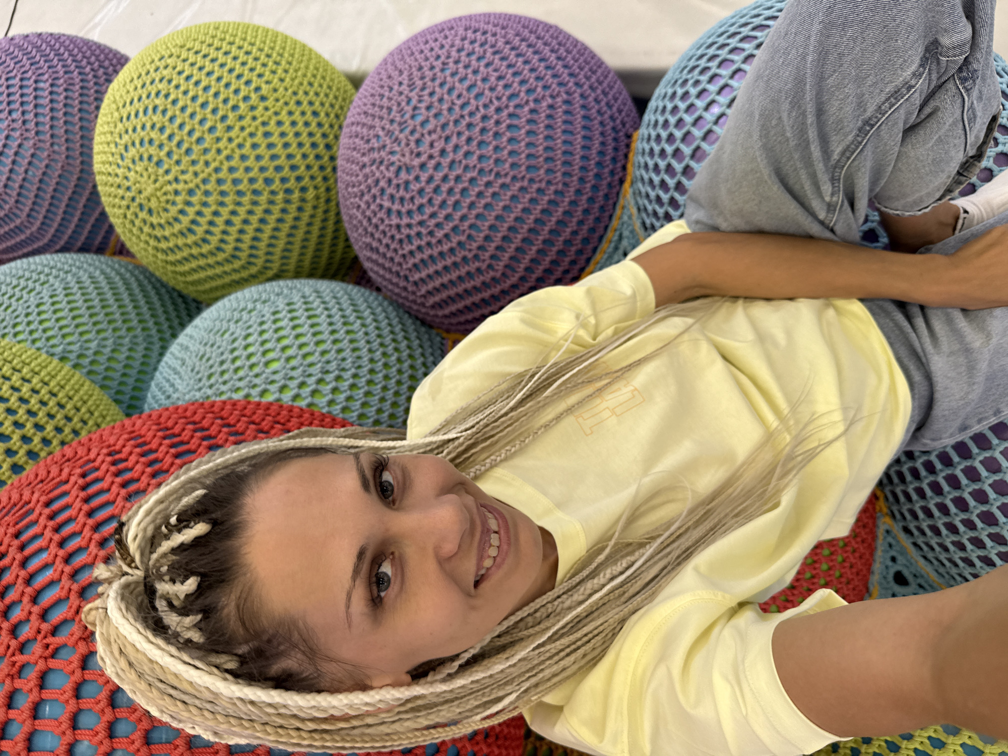 A woman with blonde braided hair smiles at the camera while sitting on large, colorful, crocheted spherical seating structures. She is wearing a light yellow t-shirt and blue jeans. The seating structures are a mix of red, blue, green, and purple. The background is out of focus but appears to be an indoor space with white walls. The location is identified as Korobovo, Russia.