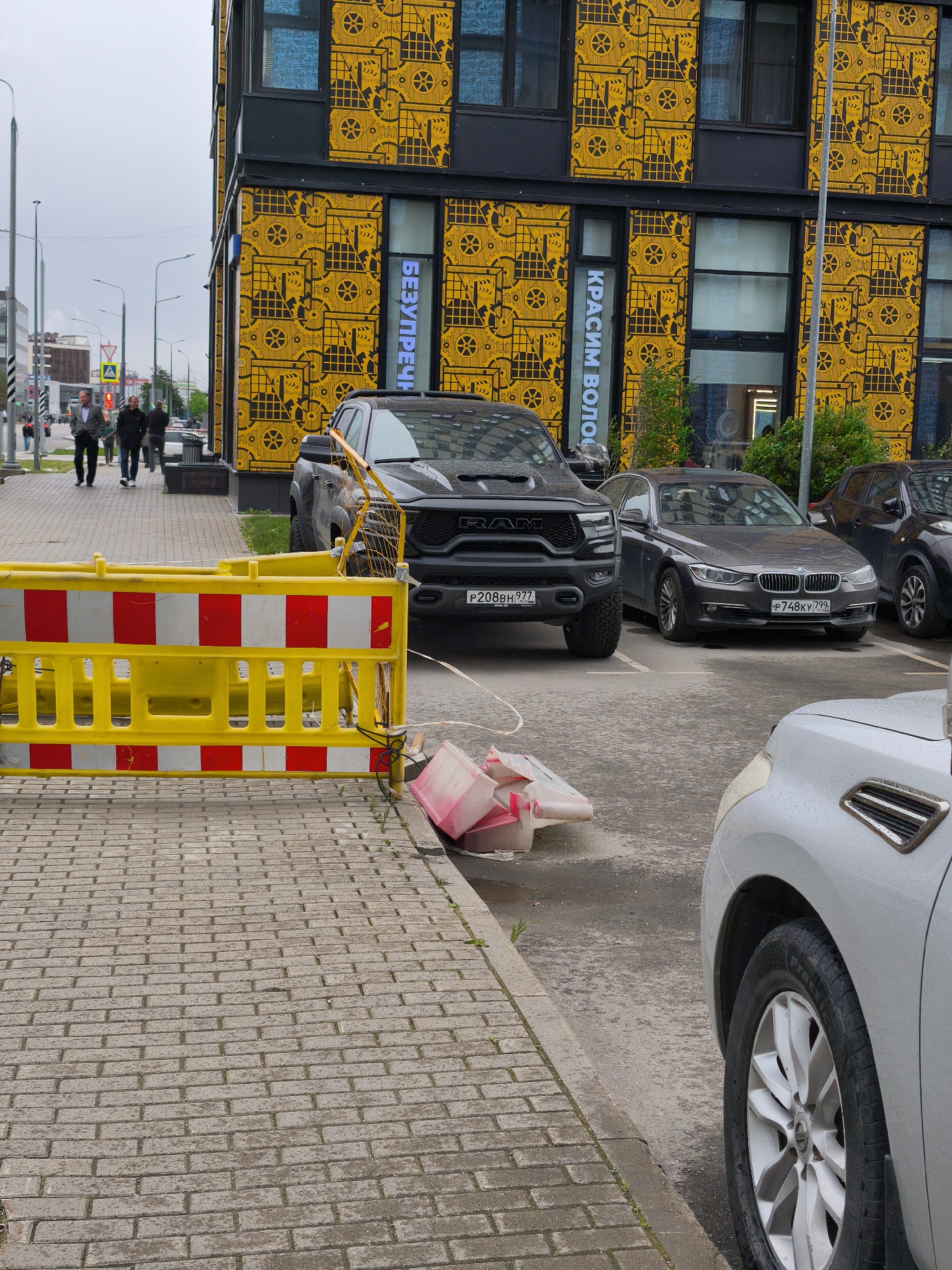 The image shows a street scene in Moscow, Russia. In the background is a building with a distinctive yellow facade decorated with black patterns resembling gears. Signs in Russian are visible on the building's facade. A yellow and white striped barrier is positioned in the foreground, partially obstructing the view.
Several cars are parked near the building, including a black RAM truck with a license plate "P208BH777", a grey BMW with the license plate "P748KY799" and a silver SUV. Debris, possibly from the barrier, is scattered on the ground. On the sidewalk in the background, two people are walking away from the camera.
The scene takes place on a paved surface next to a road. The overall atmosphere is somewhat overcast.