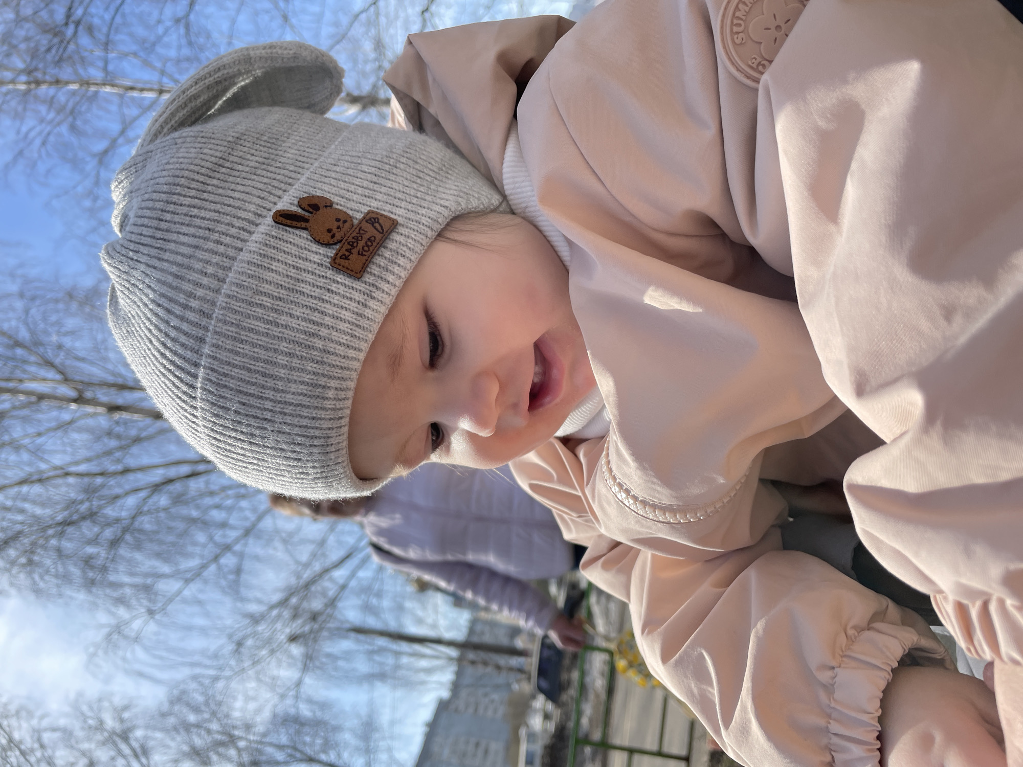 A close-up shot depicts a baby in a grey knitted hat with a "Rabbit Food" patch and a light pink jacket, positioned at an angle against a backdrop of a city park. The baby is smiling and facing downwards. The background features leafless trees against a blue sky, suggesting it is late autumn or early spring. Partially visible in the background is an adult figure wearing a light-colored jacket. The park is located in Izhevsk, Russia.