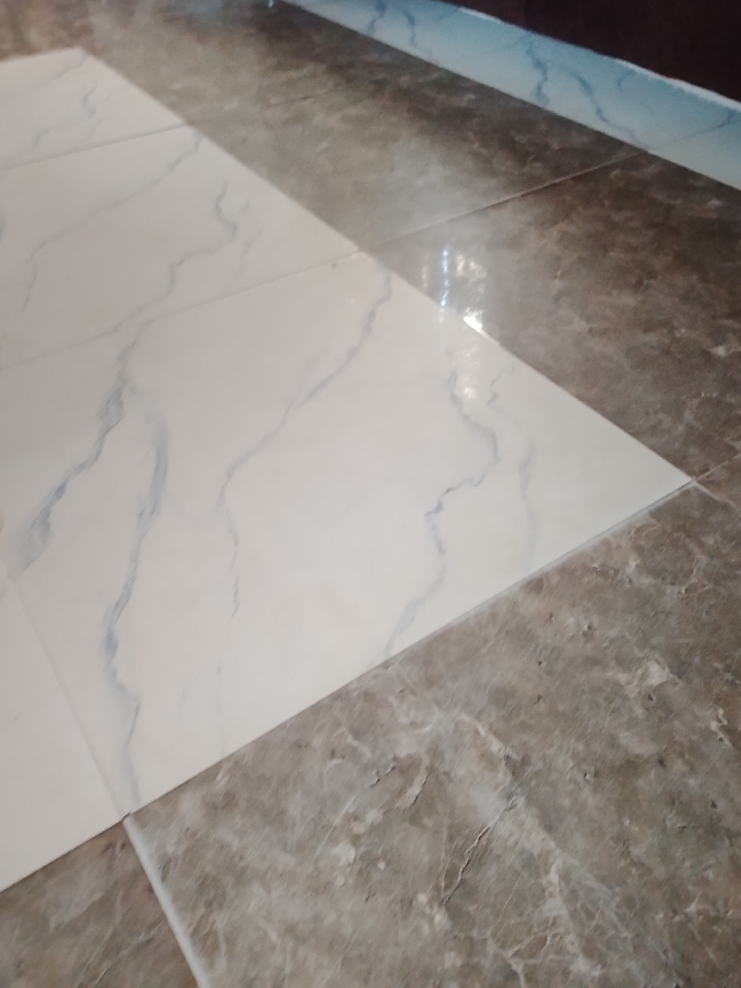 The image shows a close-up view of a tiled floor, likely in a residential or commercial building. The floor is divided into two sections with different tiling patterns. The foreground features large, white tiles with subtle blue veining, resembling marble. These tiles are laid in a diamond pattern. Behind this section, there are darker, gray tiles with a more prominent marble-like pattern. The transition between the two types of tiles is visible. The lighting suggests an indoor environment, possibly with natural light from a window or overhead illumination creating reflections on the glossy surface of the tiles. There are no people or specific objects visible that would indicate a precise location within Jalingo, Nigeria, beyond it being an interior tiled floor.