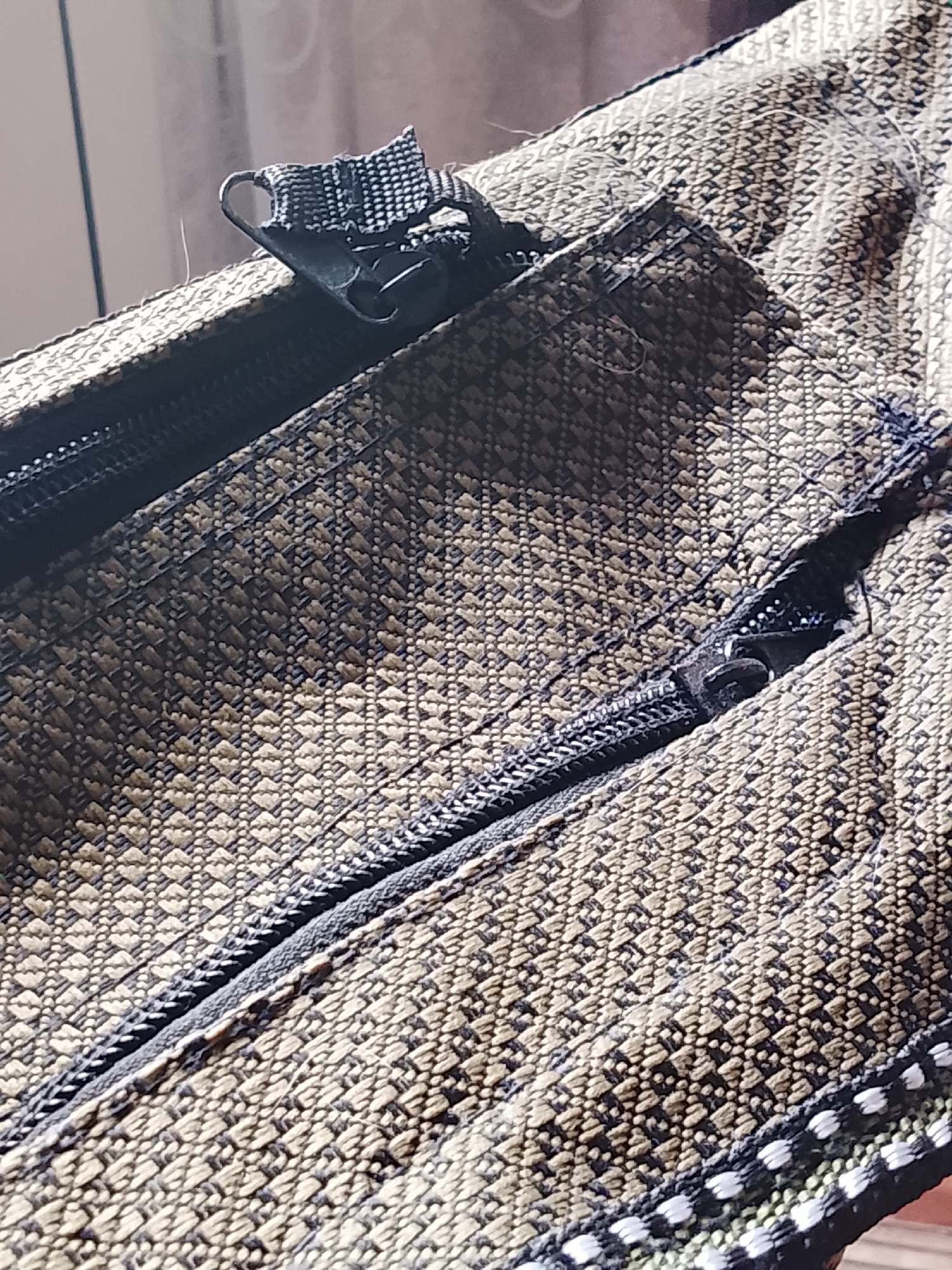 The image displays a close-up view of a brown and tan woven fabric bag with two zippers. The fabric has a textured, diamond-like pattern. One zipper is partially open, revealing the teeth of the zipper. A small black tab with a metal loop is attached to the slider of this zipper. The second zipper runs diagonally across the lower portion of the image, with its slider visible. The background is blurred, showing a light-colored curtain and a hint of a wooden surface. There are no people or animals present, and the setting is unclear, with no identifiable landmarks of Almaty, Kazakhstan.
