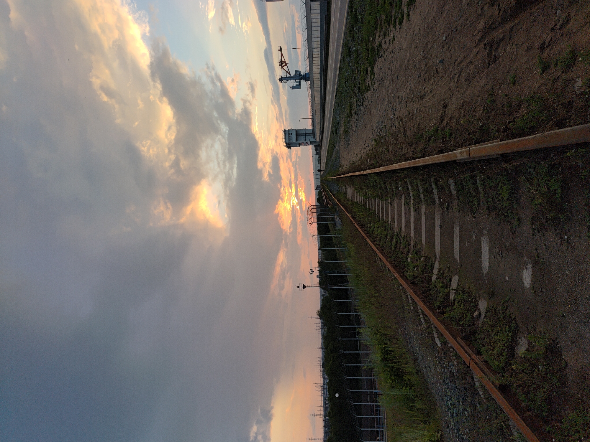 An outdoor, wide-angle, low-angle shot captures an abandoned railway track in Chaikovsky, Russia, leading towards a dramatic sunset. The sky is filled with a mix of dark, moody clouds and brightly illuminated, golden-orange clouds where the sun is setting. In the distance, on the right side of the frame, a modern concrete structure with a control tower and a large industrial crane is visible, suggesting proximity to a port or industrial area. A paved road runs parallel to the railway track on the right. The railway tracks themselves are rusty and overgrown with grass and weeds, indicating disuse. A chain-link fence topped with barbed wire runs along the edge of the industrial area. The overall impression is one of an industrial landscape juxtaposed with the natural beauty of a sunset. A single, small human figure can be seen walking on the road in the distance.