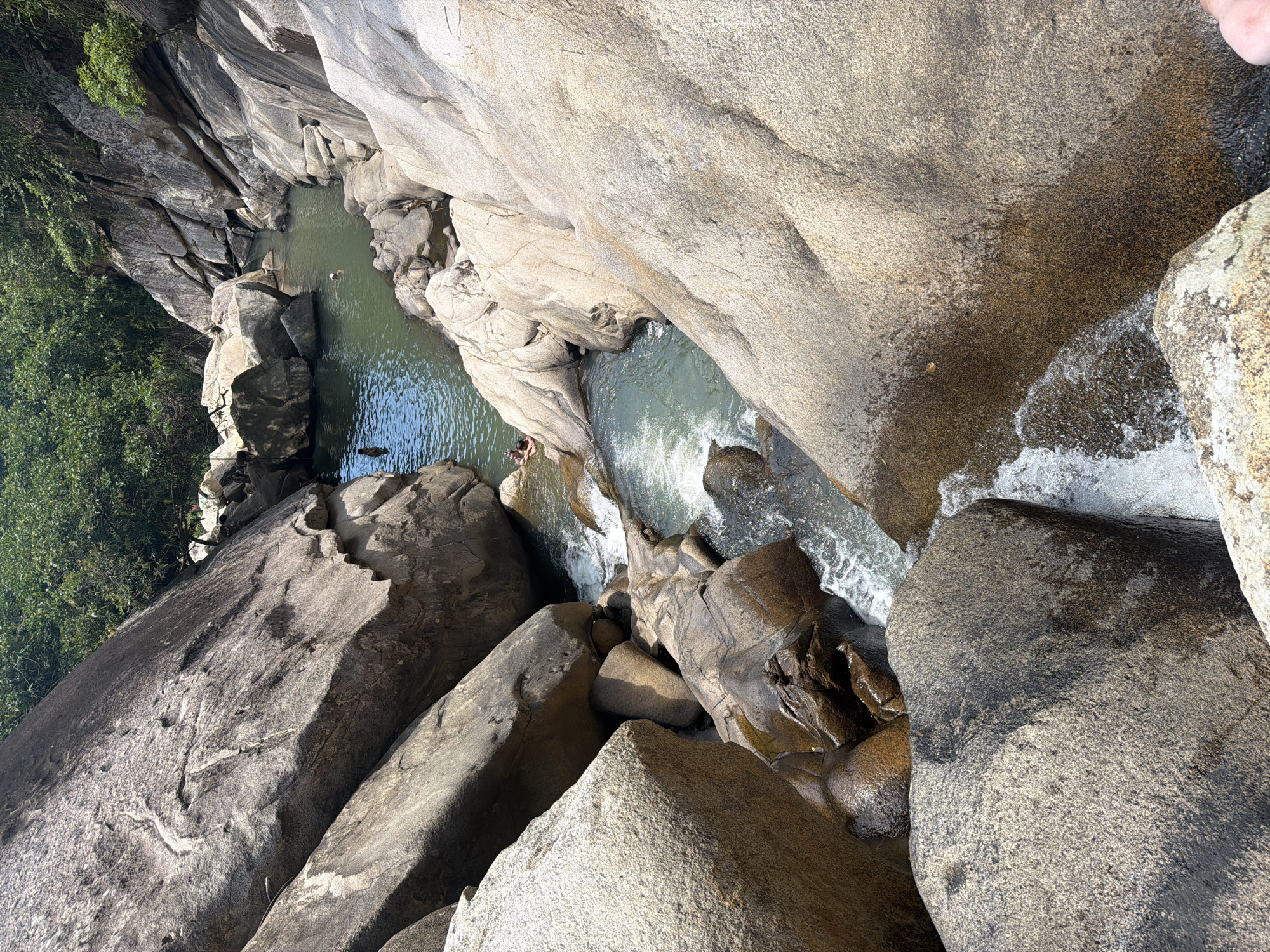 The image captures a natural rock formation at Suoi Tien, located in Hoa Thang Ward, Phan Thiết, Vietnam. The composition is an aerial view of a rocky gorge with pools of water and small waterfalls. The landscape is primarily composed of large, smooth, gray-beige rocks, creating a natural enclosure around the water. There are two discernible pools of water. In the first pool, two figures are seen swimming. The second pool is connected to the first by a small waterfall. The water is clear, with some areas appearing greenish. Some scattered vegetation is visible on the left side.