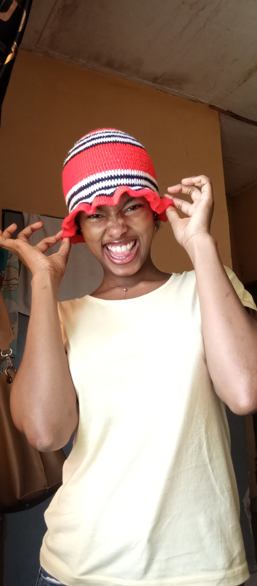 A vibrant, well-lit portrait captures a young woman indoors in Keffi, Nigeria, making a joyful and playful expression. She is the central subject, looking directly at the viewer with a wide, open-mouthed smile that crinkles her eyes and nose, revealing her teeth. She is wearing a distinctive red, white, and black striped knitted or crocheted bucket hat, which she playfully pulls down over her ears with both hands.
The woman has dark skin and is dressed in a simple light yellow, short-sleeved t-shirt. A delicate gold necklace with a small spherical pendant is visible around her neck. To the far left of the frame, parts of a brown bag strap with a gold clasp and keys are visible, possibly hanging from a hook.
The background consists of a plain, light brown or beige wall, indicating an indoor setting. The ceiling above is darker with some visible texture. The natural lighting suggests it is daytime. The woman's pose and animated facial expression convey a strong sense of happiness, amusement, and a lighthearted mood. There is no visible text in the image.