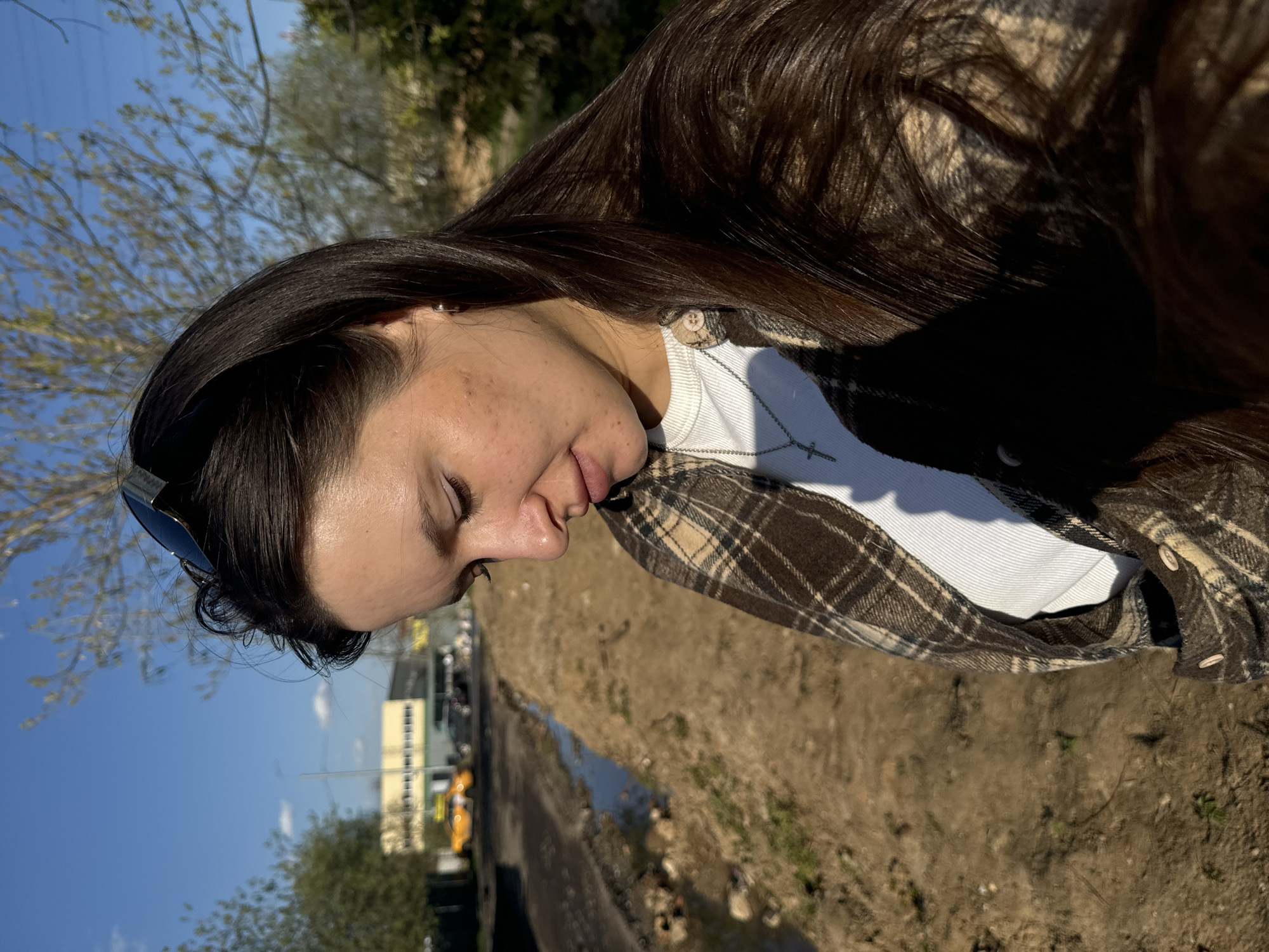 A woman with long, dark hair and closed eyes is photographed outdoors. She wears a white ribbed undershirt and a brown plaid flannel shirt. Sunglasses are perched on her head. In the background, there's a patch of bare earth with some puddles. Beyond that, a street with buildings and vehicles is visible under a blue sky. Trees with sparse foliage frame the upper and lower left corners of the image. Based on the provided location, this appears to be a scene captured in or near Akulovo, near Kubinka in Russia.