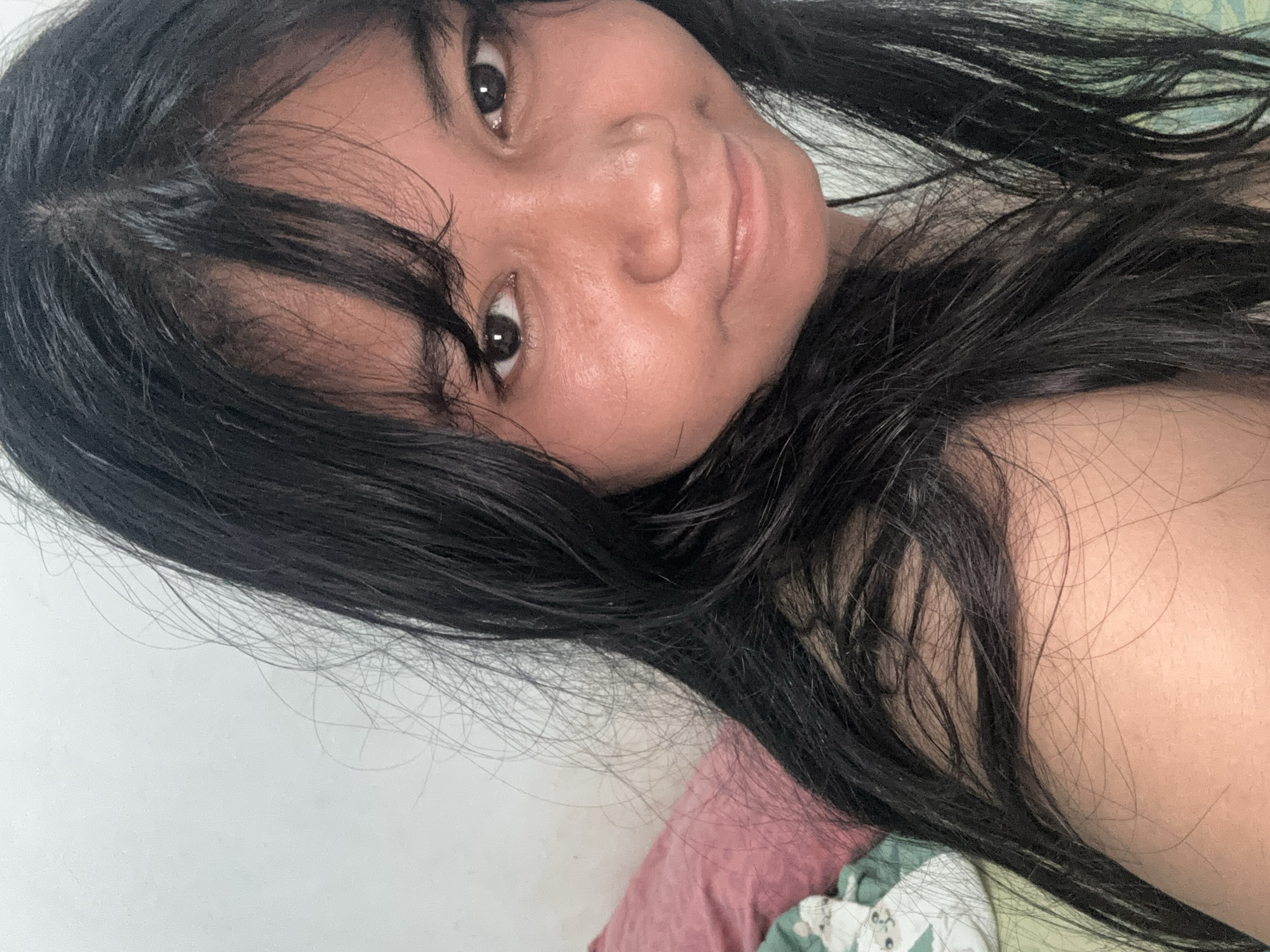 This is a close-up, eye-level selfie of a young woman with long, dark, slightly messy hair and bangs. She has dark brown eyes and appears to be looking directly at the camera with a slight smile. Her skin is a warm tone, and there are some faint blemishes or marks visible. She is positioned on a bed, with a pillow, possibly pink, and a green patterned fabric visible in the background. The lighting suggests an indoor setting, likely a bedroom, and the overall impression is casual and intimate. The scene appears to be taking place in a residential setting in North Jakarta, Indonesia, based on the provided context.