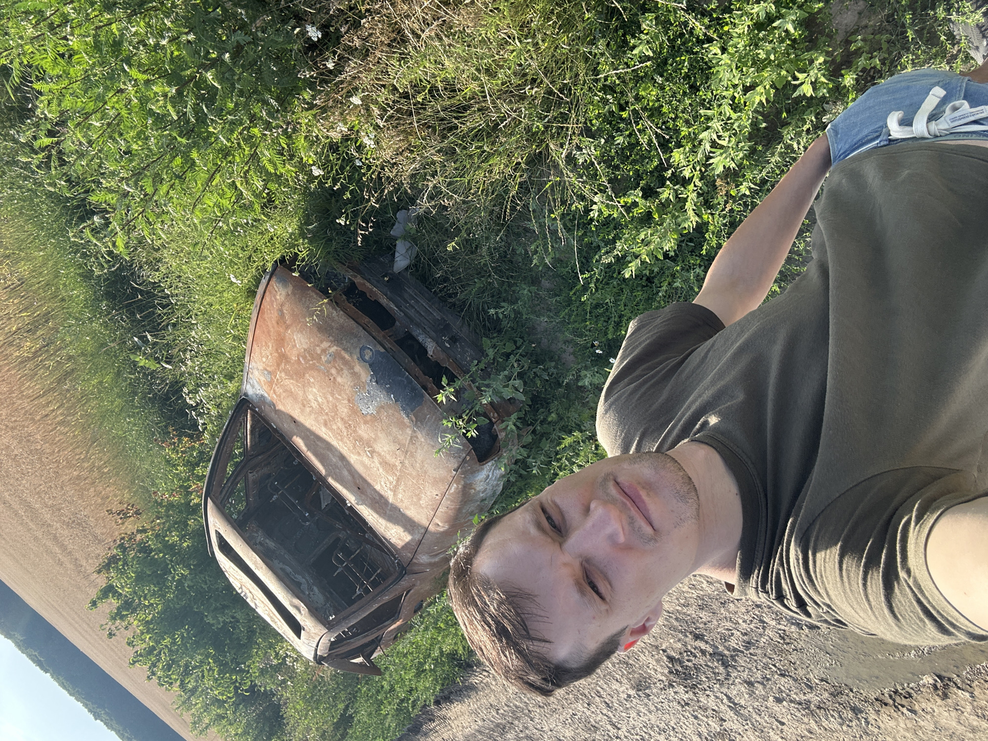 A man, wearing a dark green t-shirt and denim shorts, takes a selfie outdoors. To his right, a rusted, burnt-out car is positioned on a grassy area adjacent to a field of dry, golden crops. The ground to the left of the man is a mix of dry soil and shallow water. The environment appears to be rural, likely in Makiv, Ukraine, under clear daylight.
