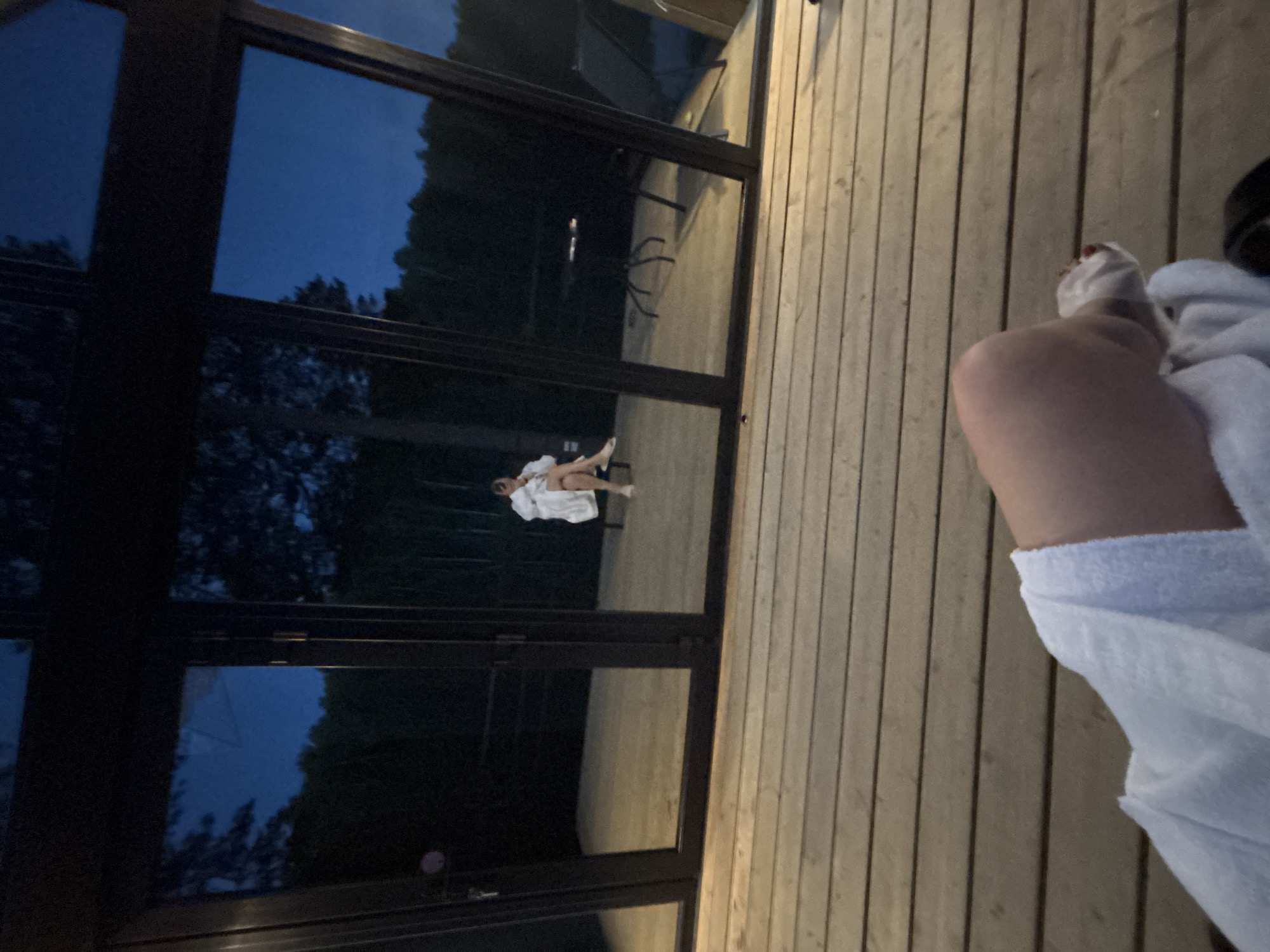The image depicts an outdoor evening scene in Mishkino, Russia, viewed from a low perspective on a wooden deck. In the immediate foreground, the lower portion of a person's body is visible, clad in a white terry cloth robe, with a foot exhibiting red painted toenails. The foreground surface is a light brown wooden deck composed of horizontal planks. The midground features a modern structure with multiple dark-framed, tall glass panels. These panels reflect a dark blue twilight sky and dark, silhouetted trees. Through the glass, a second individual is visible indoors, seated on what appears to be a light-colored bench or chair. This individual is also wearing a white terry cloth robe. Further to the right and reflected within the interior space, a dark, spindly-legged table with a few small, indistinct objects on its surface is present. The overall lighting is dim, indicative of dusk or early night, with the interior space slightly more illuminated than the exterior.
