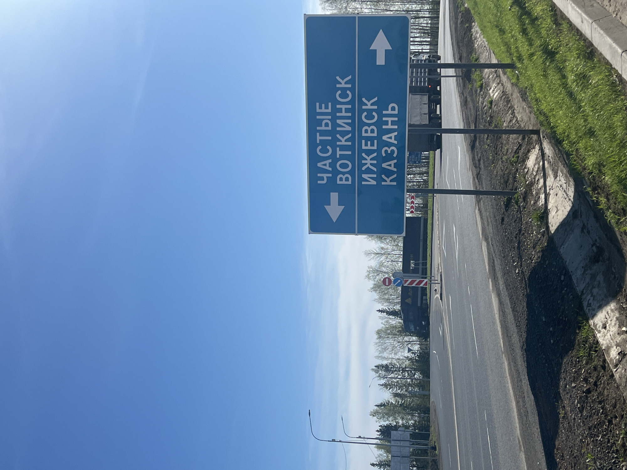 The image shows a blue road sign in Verkh-Sosnova, Russia. The sign has white lettering in Russian, with arrows pointing left and right. The left arrow is next to the words "ЧАСТЫЕ ВОТКИНСК" and the right arrow is next to the words "ИЖЕВСК КАЗАНЬ". The sign is mounted on metal poles next to a gray asphalt road. There is green grass and vegetation alongside the road. In the background are trees and a clear blue sky. A truck is partially visible behind the sign.