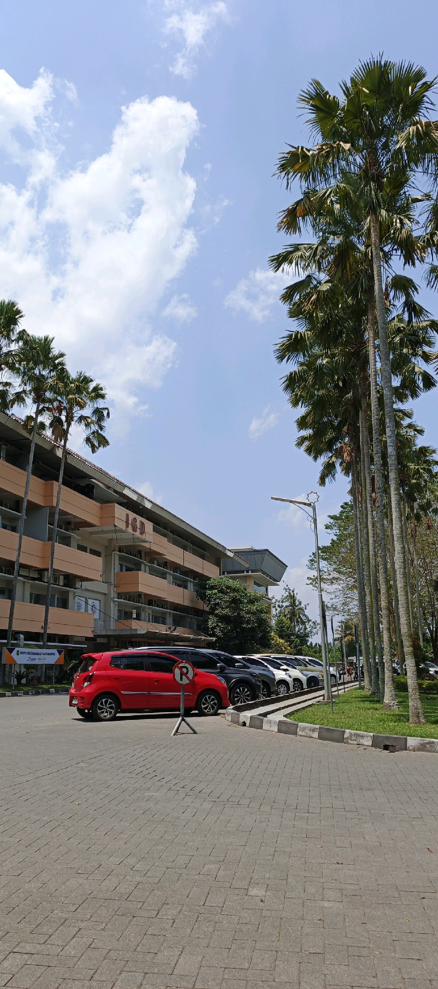 The image depicts a scene in front of a large building, likely a hospital or medical facility, located in Ambarketawang, Indonesia. The building, which appears to be several stories high with an orange facade, has "IGD" visible on its upper level, indicating an Emergency Department. In the foreground, a red car is parked, accompanied by a "no parking" sign. Several other cars are lined up in a parking lot. Tall palm trees are prominent in the scene, lining the parking area and the building's exterior. The sky is blue with scattered white clouds. The ground is paved with interlocking concrete blocks. The overall impression is an outdoor view of a public facility on a clear, sunny day.