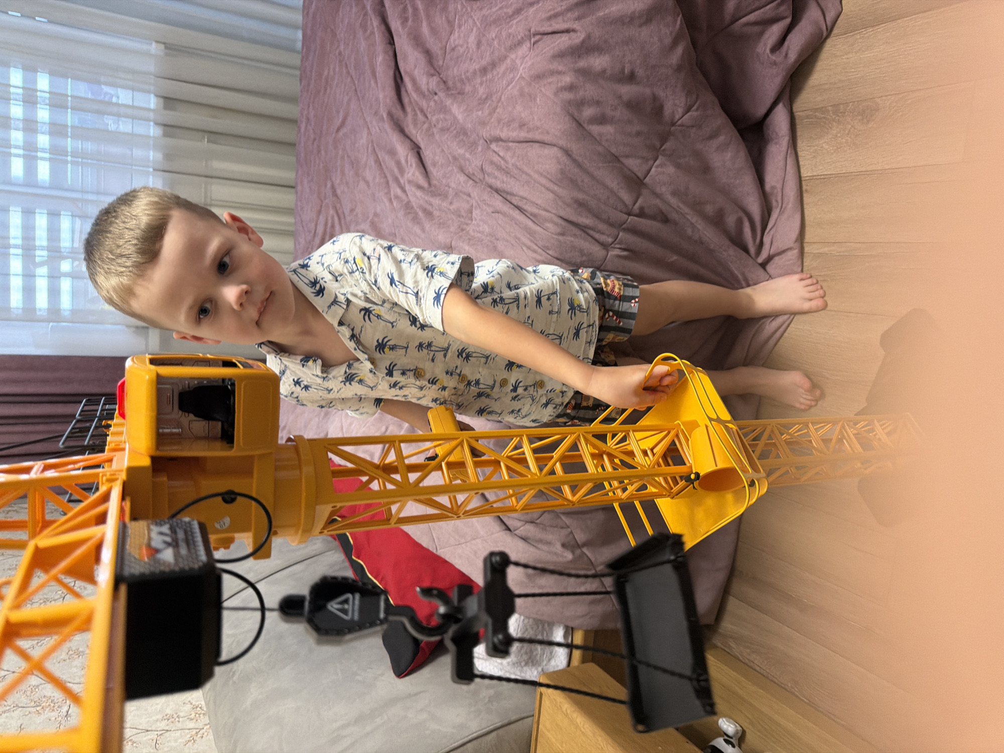 In this indoor scene, a young boy with blond hair is lying on a bed next to a large yellow toy crane. He is wearing a short-sleeved shirt with a palm tree pattern and shorts. The boy is looking towards the toy crane, with his head tilted and his expression suggesting engagement or curiosity. His right arm is extended, and his hand is near the crane's controls, as if he is playing with it.
The bed has a rumpled, dark purple comforter. The floor next to the bed is made of light brown wood planks. In the background, sheer white blinds cover a window, and a dark pink curtain is visible.
The toy crane is bright yellow and has a detailed structure, including a cabin with black interior details and a boom arm. A black cable hangs from the boom, attached to a hook. Other toy construction equipment, including what appears to be a black and red toy excavator, are also visible on the bed.
The lighting suggests it could be daytime, possibly afternoon, given the soft light filtering through the blinds. The overall atmosphere is relaxed and playful, indicative of a child at home. No visible text is present in the image. The location context is Minsk, Belarus.