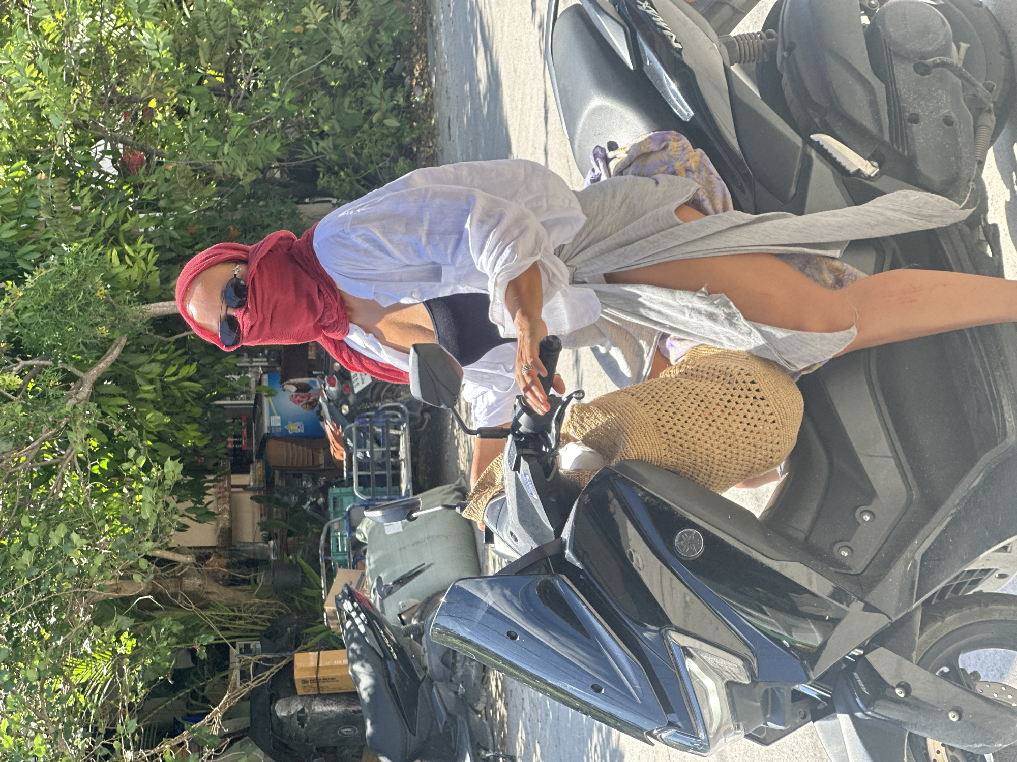 A woman with a red headscarf and sunglasses is sitting on a black Yamaha motorcycle in what appears to be a cluttered area in ตำบลเกาะพะงัน, Thailand. She wears a white shirt and a black top underneath. The motorcycle is parked on a paved surface, with a straw bag and other items placed on the seat. The background contains lush greenery, and a glimpse of a building with assorted items cluttered around it.