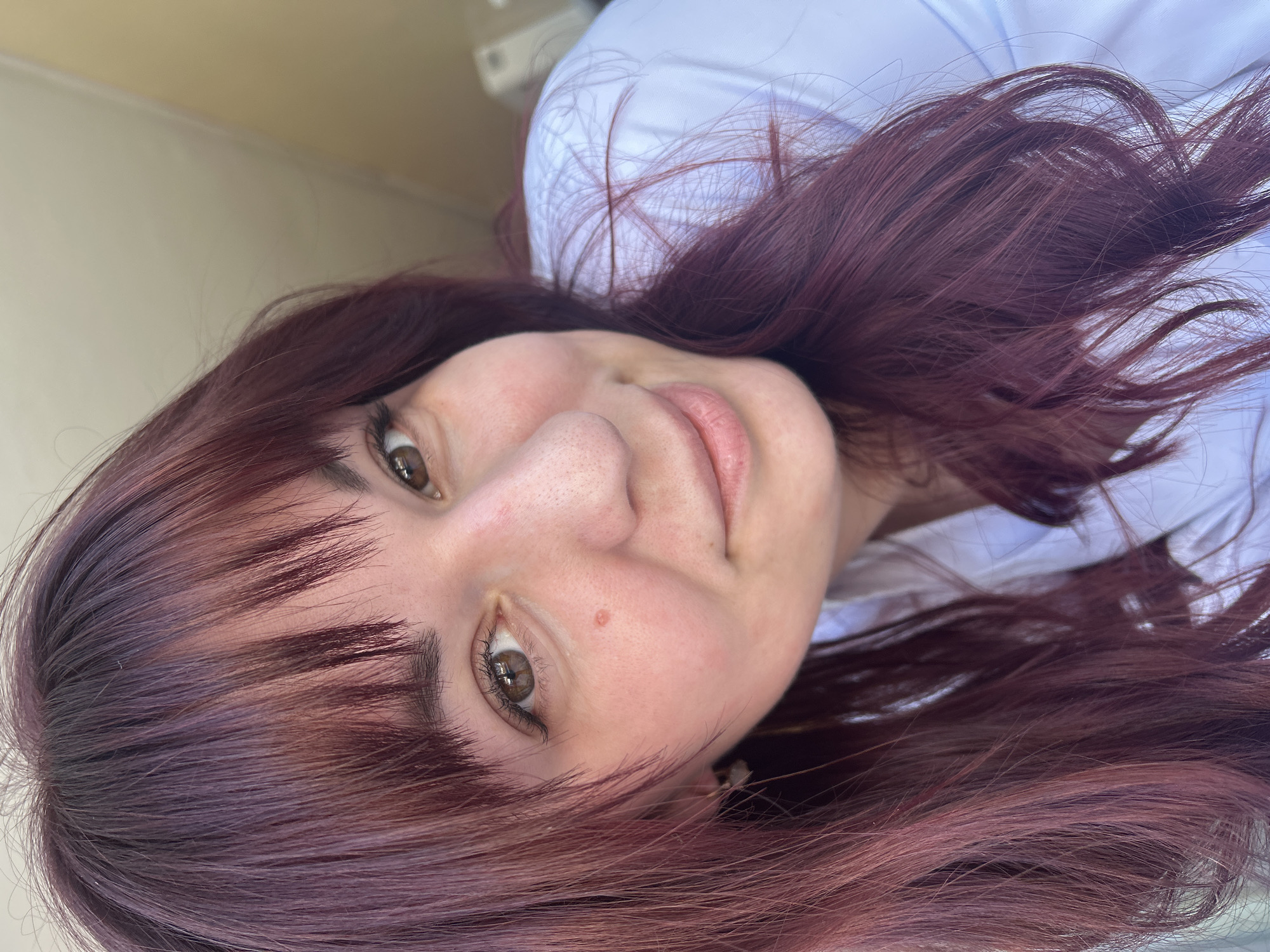 A close-up headshot of a woman with burgundy hair and bangs. She has fair skin, light brown eyes, and a mole on her left cheek. She is wearing a white shirt. The background is a light beige or cream color, suggesting an interior setting. The location is claimed to be in the city of Фоки, Russia, although this cannot be confirmed from the image alone.