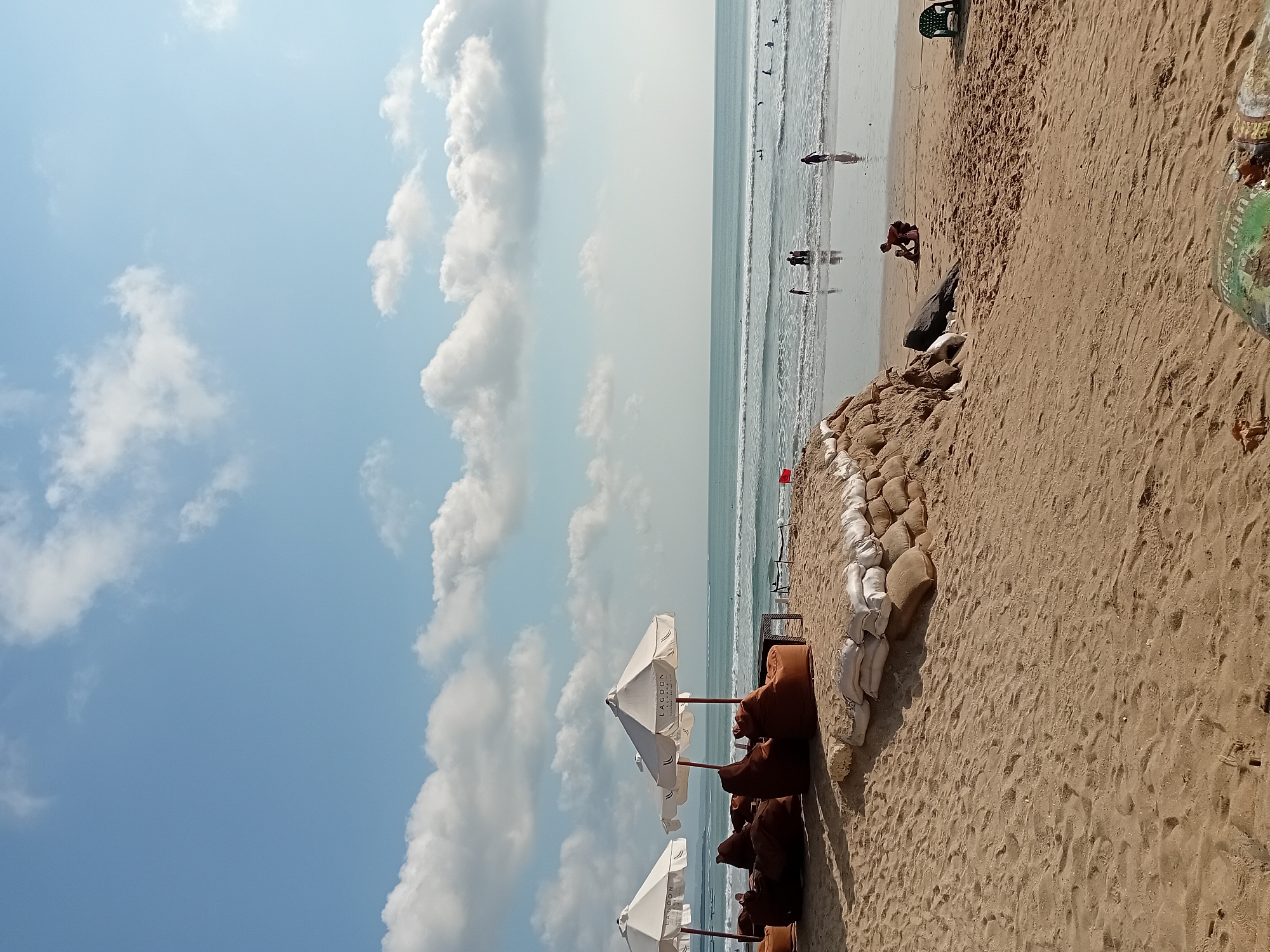 The image depicts a sunny day at Kuta Beach, Bali, Indonesia, with a clear blue sky dotted with white, fluffy clouds. The foreground is a sandy beach with footprints and some debris. In the mid-ground, there is a raised sand embankment fortified with sandbags, and to the left, there are several large, brown beanbag chairs and two white beach umbrellas, one of which has "LAGOOON GOURMET" printed on it. Beyond this seating area, the ocean stretches out, with gentle waves rolling onto the shore. Several people are visible in the water, some appearing to be surfers or bodyboarders. A few individuals are also standing and walking in the shallow water near the beach. To the right of the sandbags, a child is playing in the sand, and a green plastic chair is placed further down the beach. The overall atmosphere is one of a relaxed beach scene.