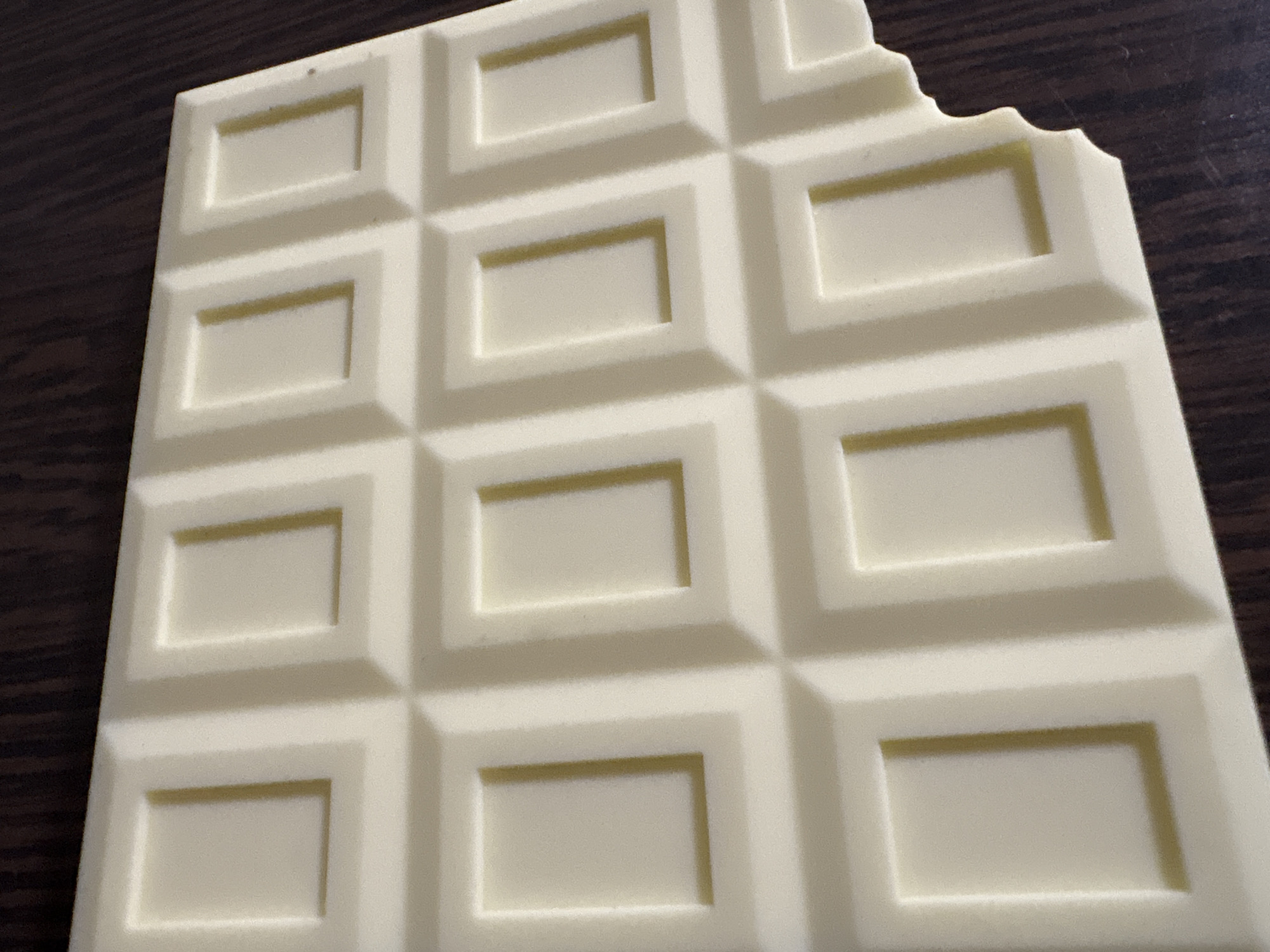 A close-up shot of a bar of white chocolate with a bite taken out of the top right corner. The chocolate is segmented into rectangular blocks. The surface underneath appears to be a dark brown wooden table. There is no indication of location in Chaikovsky, Russia in the image.