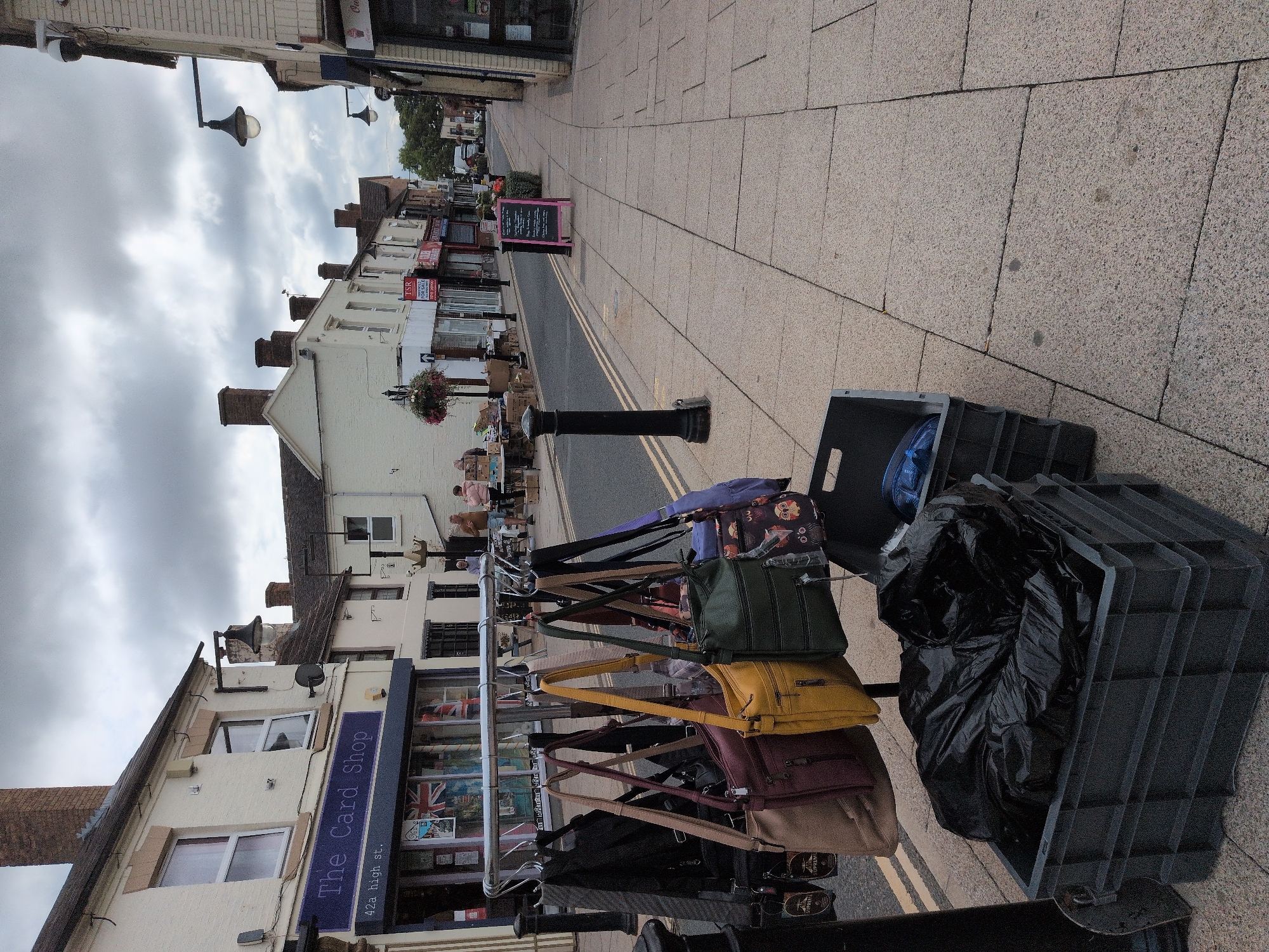 The image shows a street scene in Telford, United Kingdom. The foreground features a metal rack displaying various handbags, with some bags also in a dark grey plastic crate filled with black plastic bags. Behind the handbags, the street is lined with shops. One shop sign reads "The Card Shop 42a high st.". Further down the street, more shops are visible, along with pedestrians and parked vehicles. The sky is overcast.