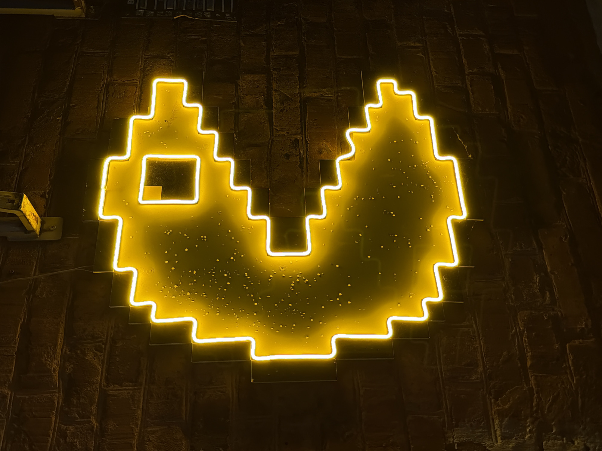 A bright, pixelated yellow neon sign, strongly resembling the classic video game character Pac-Man with an open mouth and a square eye, is prominently displayed against a dark, textured wall. The sign is formed by glowing yellow neon tubes, casting a warm, luminous aura onto the rugged surface behind it, which appears to be brick or stone. Small, scattered droplets or specks are visible on the clear backing of the neon sign.
The scene suggests an indoor setting, likely an entertainment venue, bar, or arcade, where the retro-themed signage serves as a focal point. The ambient lighting is dim, emphasizing the vivid glow of the neon. The overall atmosphere evokes nostalgia for vintage gaming and a sense of playful, inviting warmth. There are no people or specific activities depicted. The time of day is implied to be evening or night due to the sign being the primary light source in the visible area. No discernible text is present in the image. The location context is Chaikovsky, Russia.
