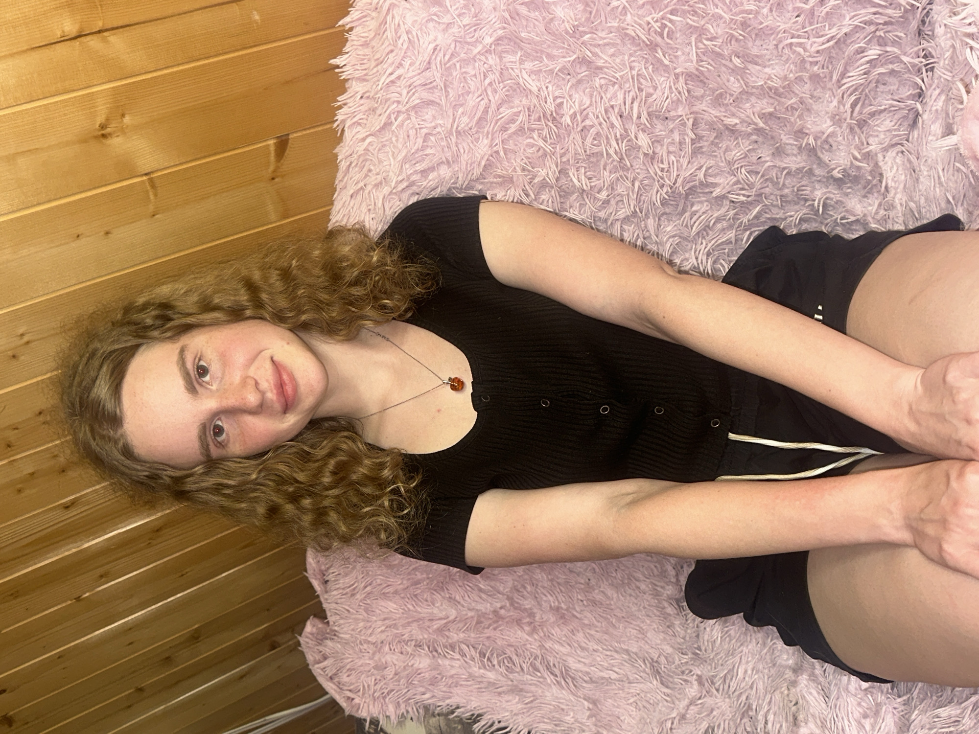 A young woman with long, wavy brown hair and a fair complexion lies diagonally across a plush, light purple, shaggy blanket, looking at the camera with a gentle smile. She has warm-toned eyes, possibly with a reddish tint, and subtle freckles visible across her nose and cheeks. She is dressed in a fitted black ribbed short-sleeved top with small button details down the front, and black shorts with white piping or drawstrings along the sides. A silver necklace with a reddish-orange, possibly amber, pendant hangs around her neck. Her arms are bent, with her hands clasped together near her lap. To the upper left of the frame, a wooden-paneled wall forms the background, indicating an indoor setting. The overall impression is one of comfort and relaxation, capturing a casual pose in Vasilyevo, Russia, likely within a home environment. There is no visible text.